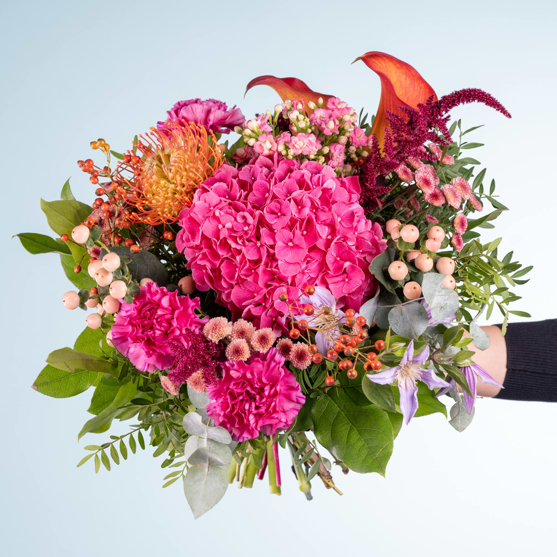 Ein lebendiger Strauß aus rosa Hortensien, orangefarbenen Dahlien und anderen bunten Blumen vor einem schlichten blauen Hintergrund.
