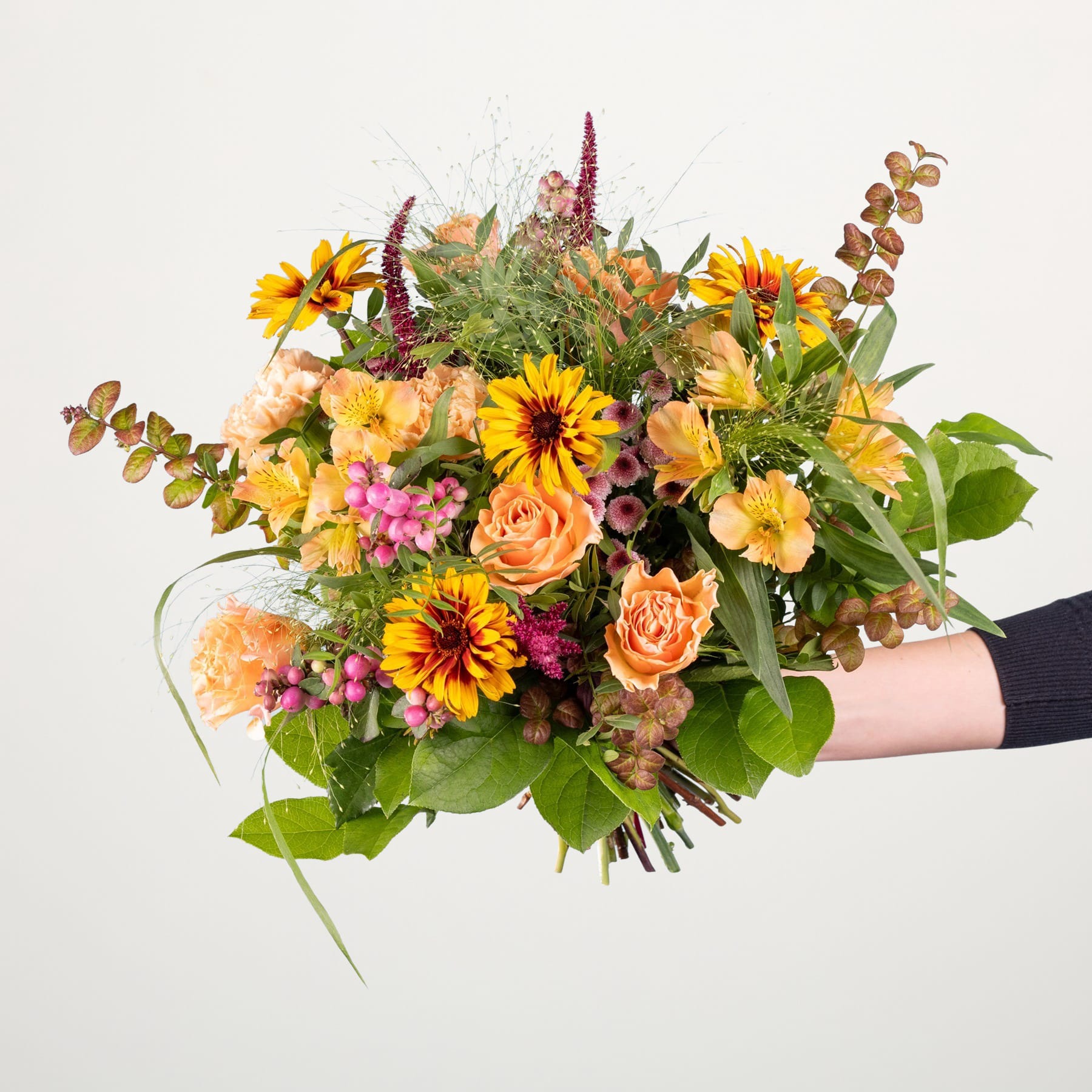 Ein lebendiger Strauß bunter Herbstblumen, darunter Sonnenblumen, Dahlien und Rosen, die eine Person vor einem schlichten weißen Hintergrund in den Händen hält.