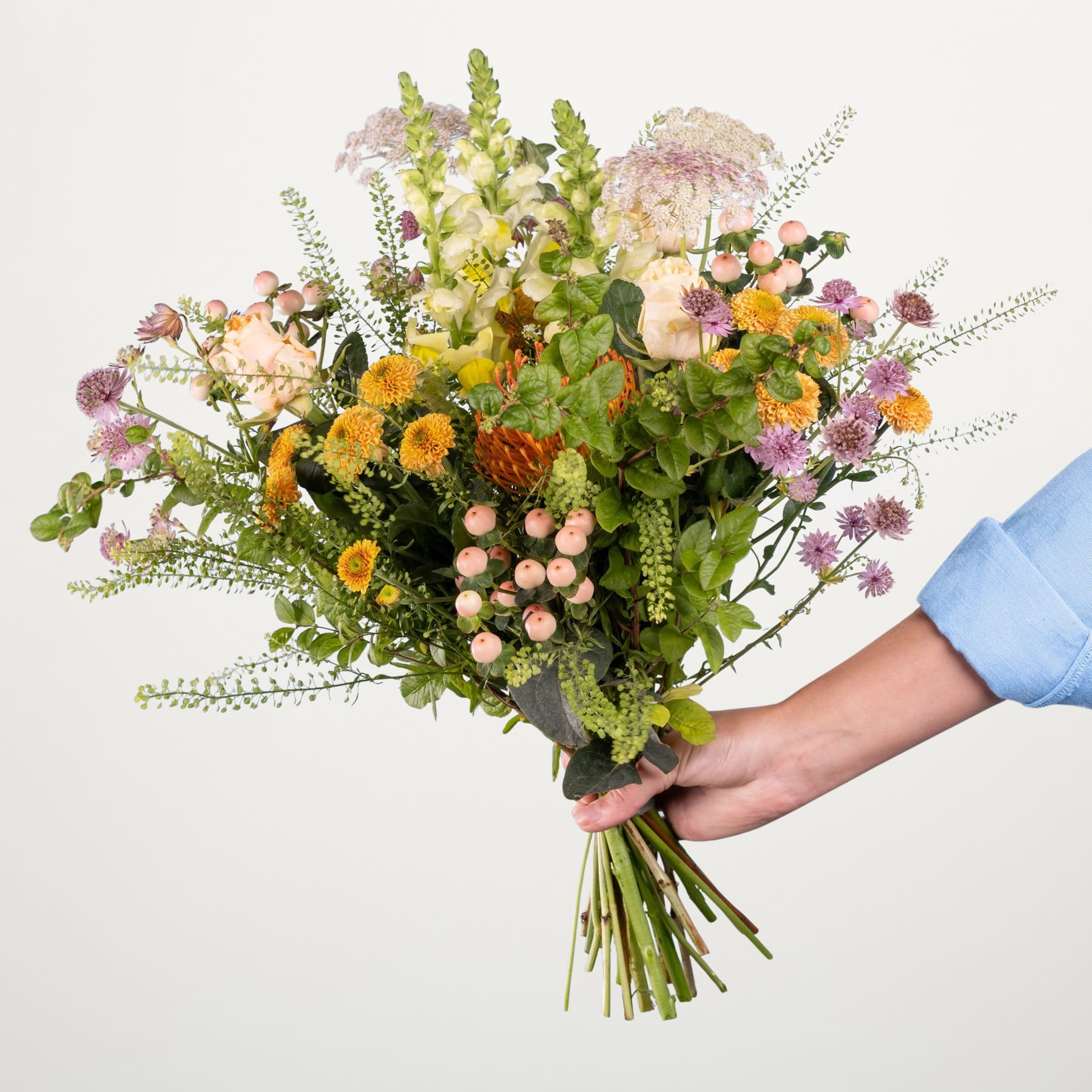 Ein leuchtender Strauß verschiedener bunter Blumen, darunter Gänseblümchen, Chrysanthemen und andere Blüten, den eine Person vor einem schlichten weißen Hintergrund in den Händen hält.