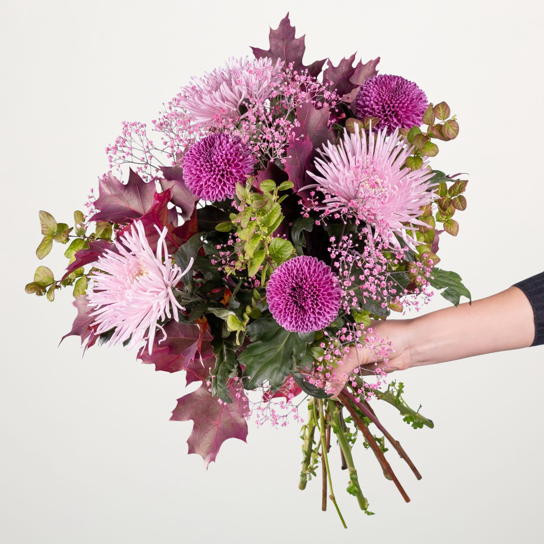 Ein leuchtender Strauß rosa und violetter Blumen, darunter Chrysanthemen und Ahornblätter, wird vor einem schlichten Hintergrund in den Händen einer Person gehalten.