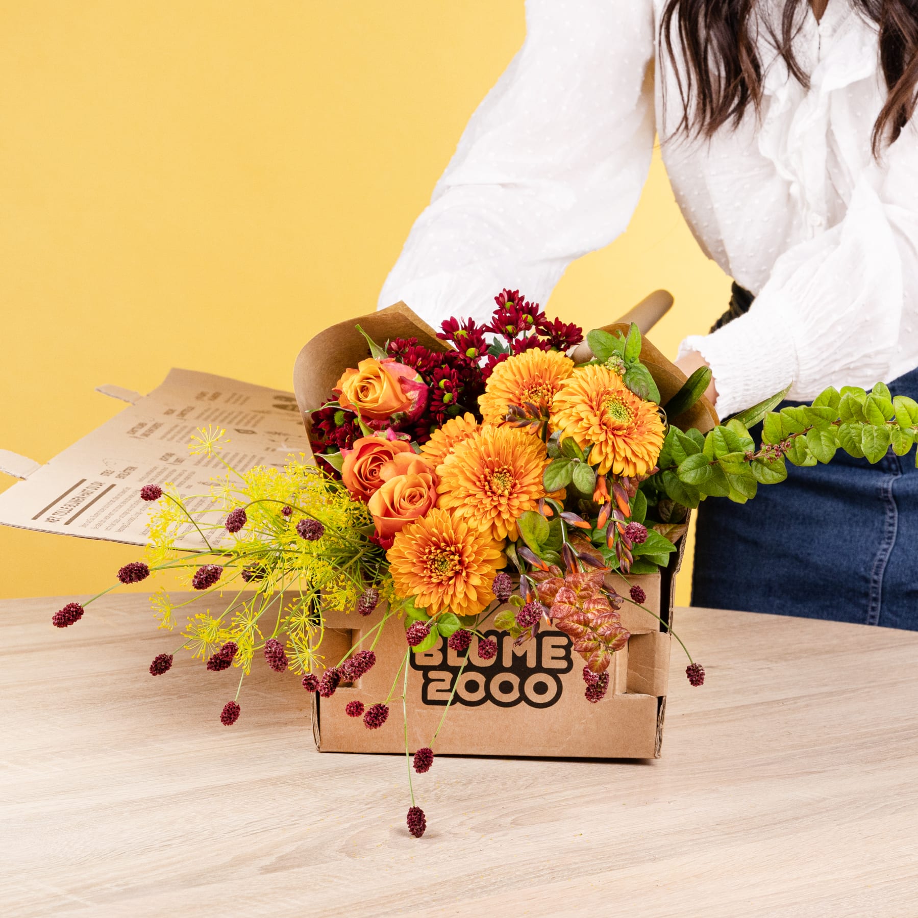 Ein farbenfroher Strauß orangefarbener und roter Blumen, darunter Dahlien und Beeren, steht in einem Karton auf einer Holzoberfläche vor einem gelben Hintergrund.