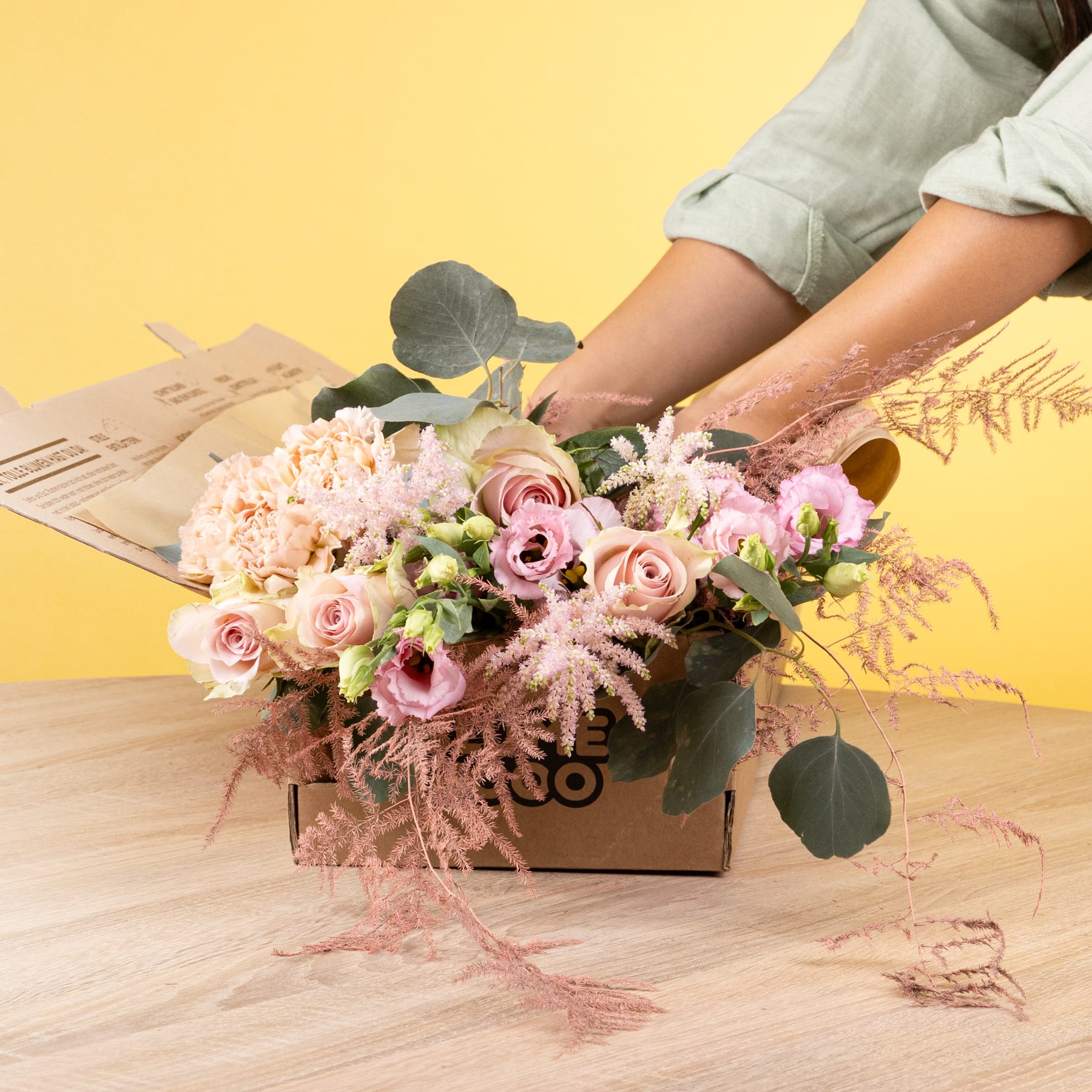 Eine Holzkiste mit einem farbenfrohen Arrangement aus rosa Rosen, weißen Blumen und Grünpflanzen steht auf einer Holzoberfläche vor einem schlichten gelben Hintergrund.