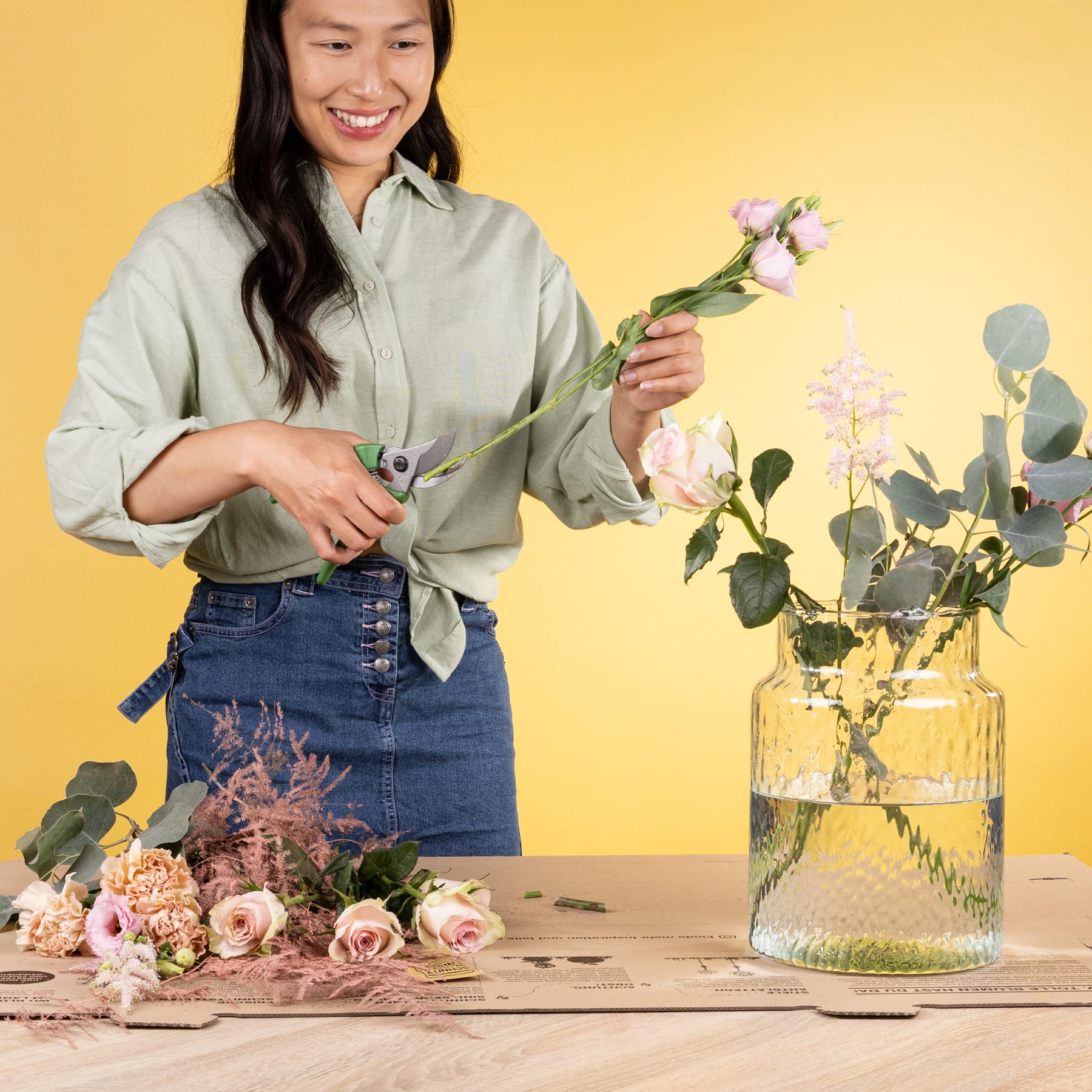 Eine junge Frau mit langem dunklem Haar arrangiert einen Strauß rosa und weißer Blumen auf einem Holztisch vor einem leuchtend gelben Hintergrund.