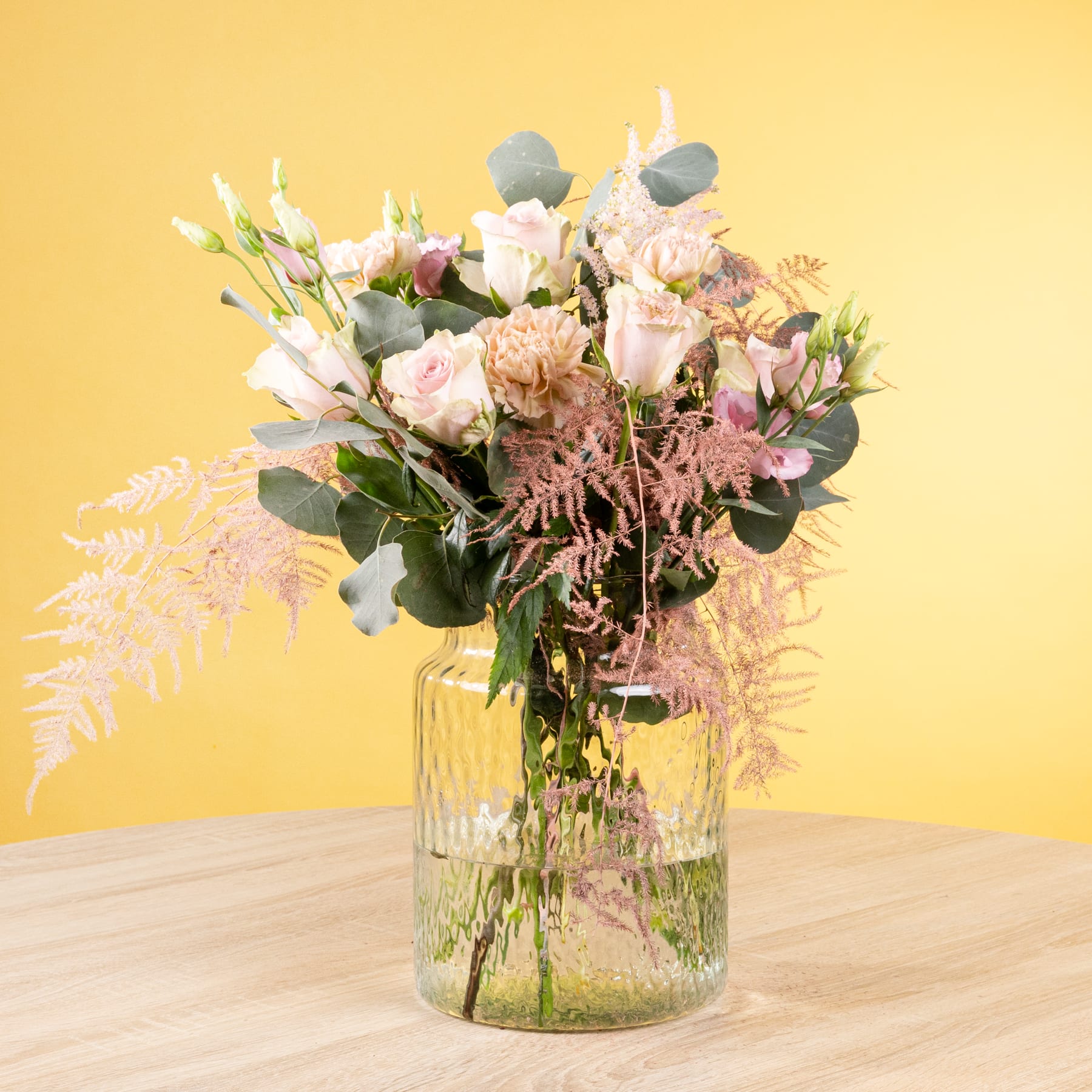 Ein lebendiger Strauß zartrosa und weißer Blumen, darunter Rosen und Farne, wird in einer Glasvase vor einem warmen gelben Hintergrund präsentiert.