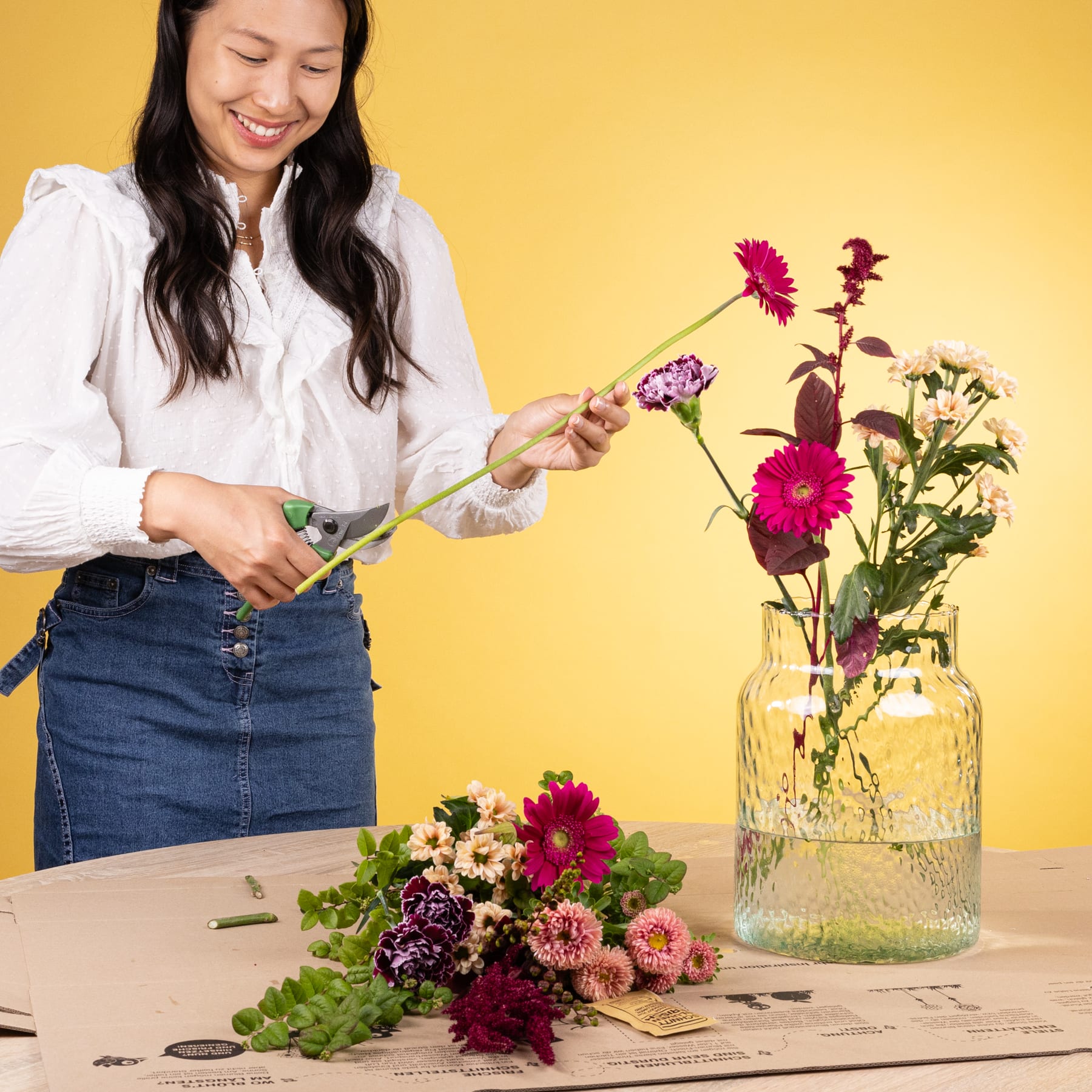 Eine junge Frau mit langem, dunklem Haar arrangiert einen Strauß bunter Blumen auf einem Tisch vor einem leuchtend gelben Hintergrund.