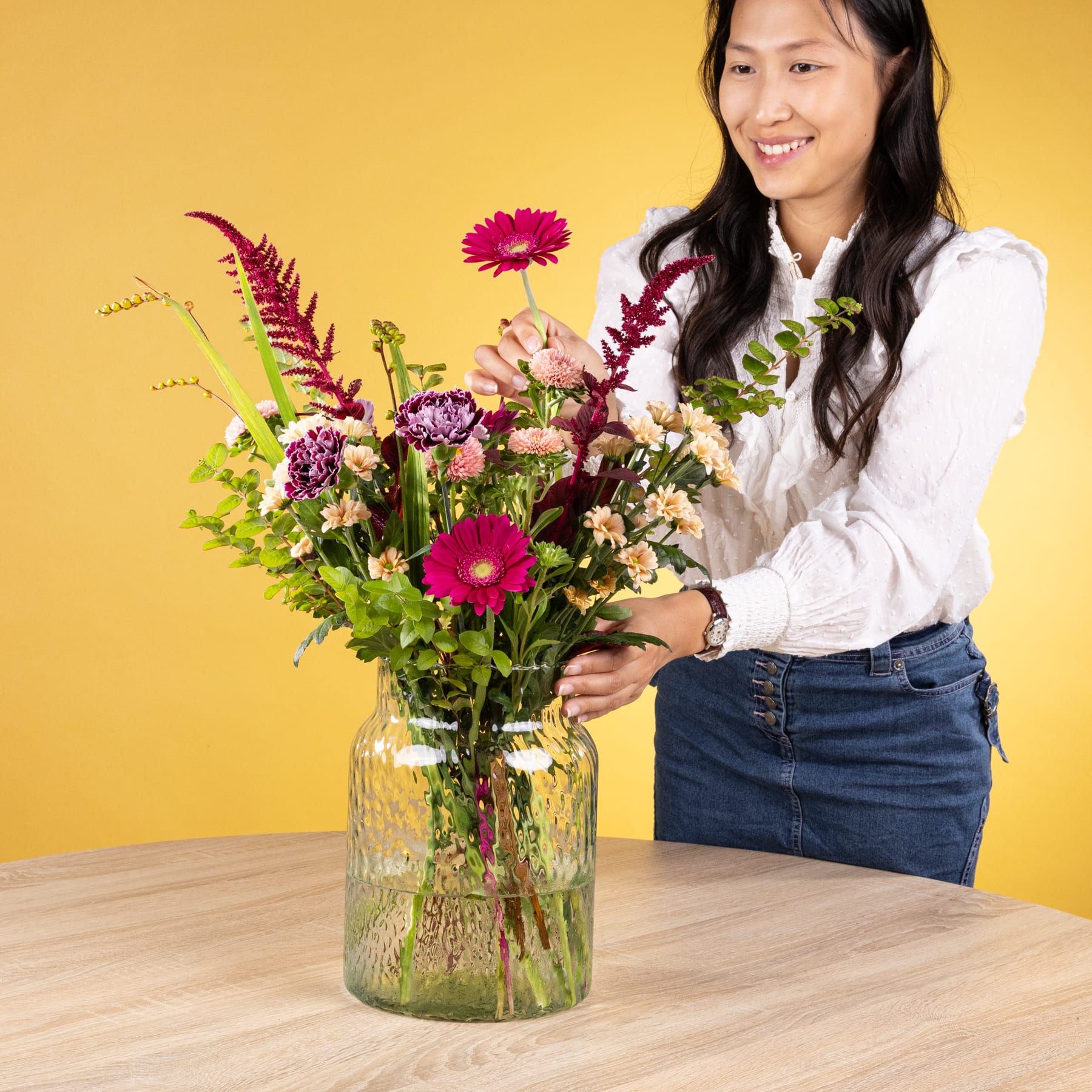 Eine junge Frau mit langem, dunklem Haar hält vor einem leuchtend gelben Hintergrund einen farbenfrohen Strauß bunter Blumen in der Hand, darunter rote Gerbera und violette Blumen.