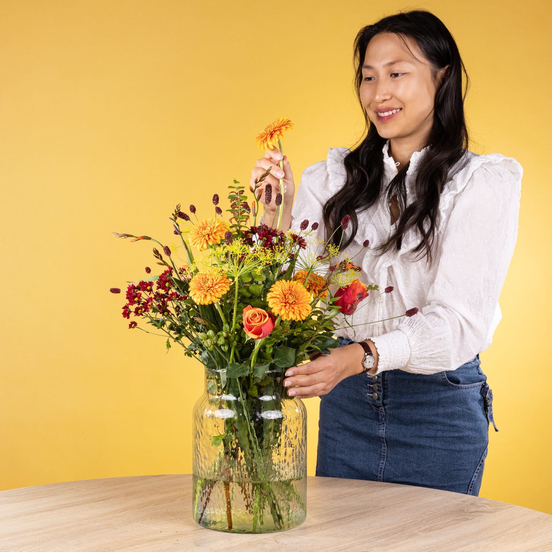 Eine junge Frau mit langem, dunklem Haar hält eine Vase mit einem farbenfrohen Blumenstrauß in der Hand und steht vor einem leuchtend gelben Hintergrund.