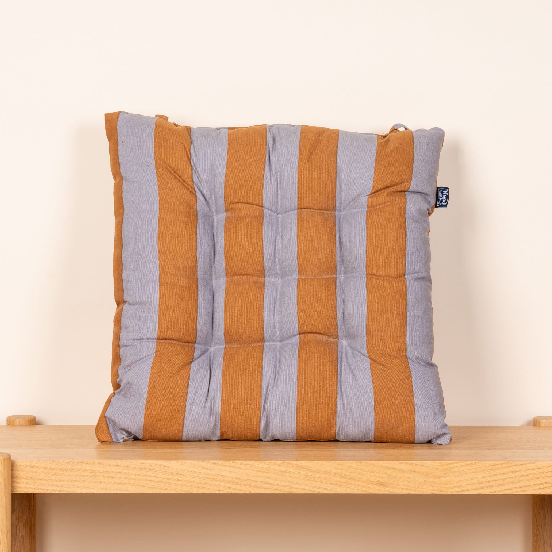 Ein orange-grau gestreiftes Kissen liegt auf einem Holzregal vor einer schlichten weißen Wand.