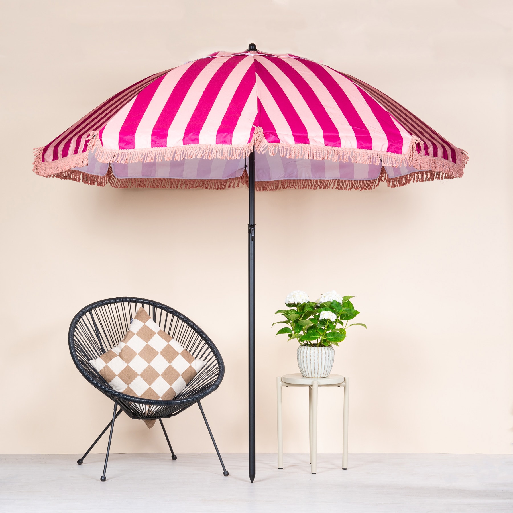 Ein leuchtend rosa und weiß gestreifter Regenschirm steht über einem geflochtenen schwarzen Stuhl und einer Topfpflanze auf einer weißen Fläche vor einem schlichten Hintergrund.
