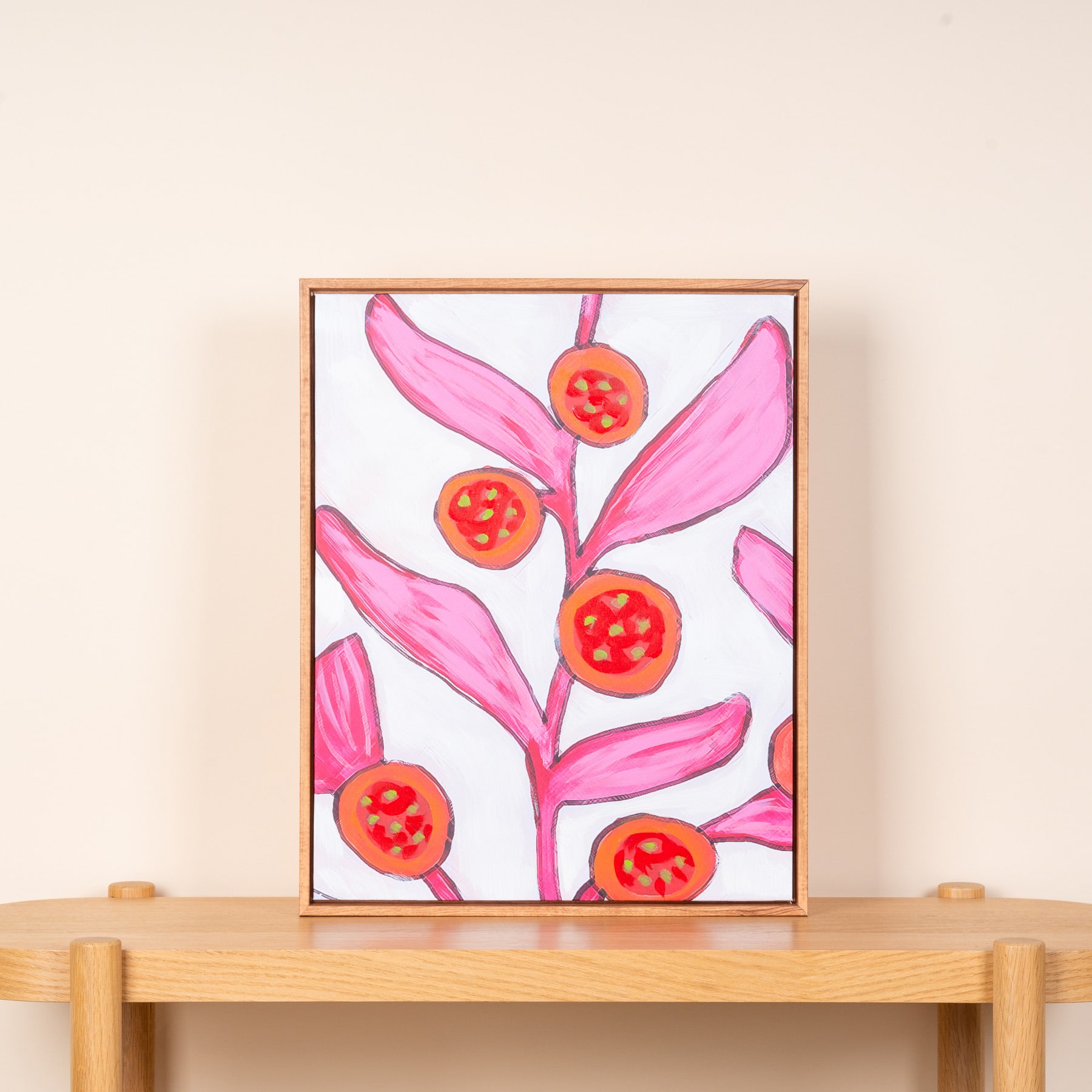 Das Bild zeigt ein lebendiges Blumenarrangement mit großen rosa Blättern und leuchtend roten Blüten oder Beeren vor einem schlichten, hellen Hintergrund. Das Kunstwerk ist gerahmt und wird auf einem Holzregal ausgestellt.