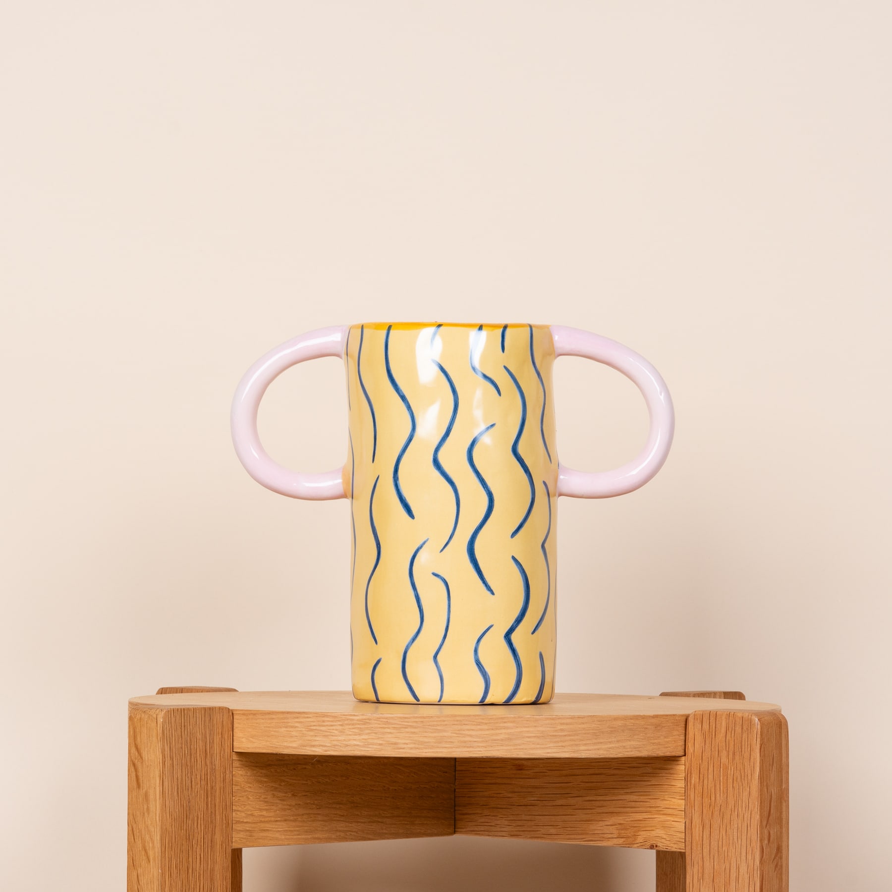Eine gelbe Keramikvase mit Wellenmuster steht auf einem Holzständer vor einem schlichten beigen Hintergrund.