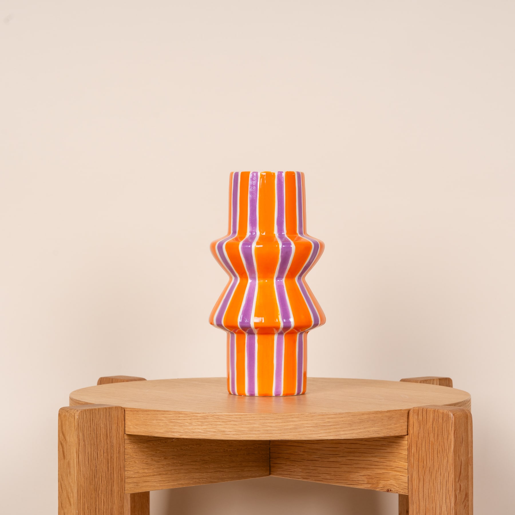Eine farbenfrohe, gestreifte Keramikvase steht auf einem Holzsockel vor einem neutralen beigen Hintergrund.