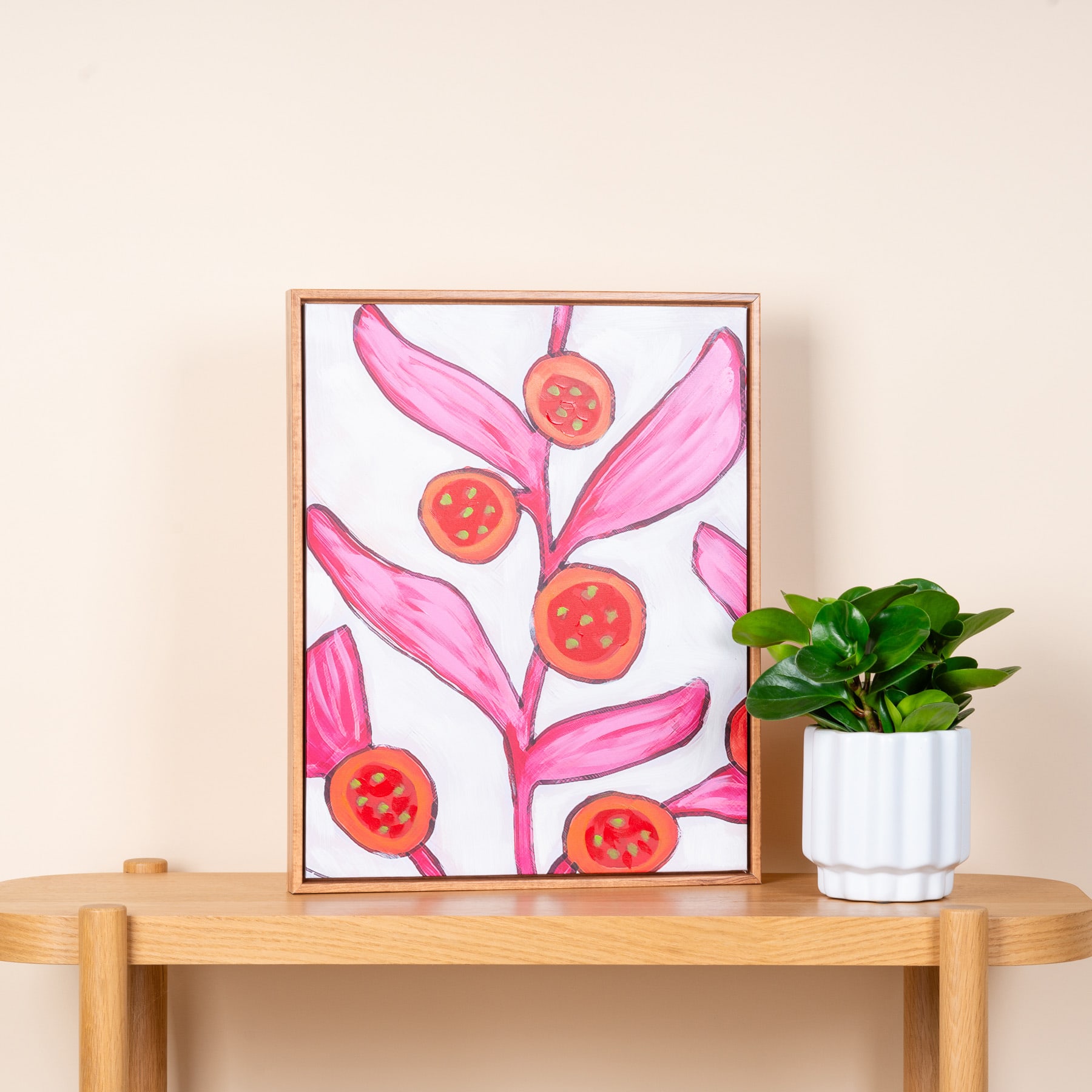 Ein gerahmtes Kunstwerk mit leuchtend rosa Blättern oder Blütenblättern mit roten kreisförmigen Formen, platziert auf einem Holztisch neben einer Topfpflanze.