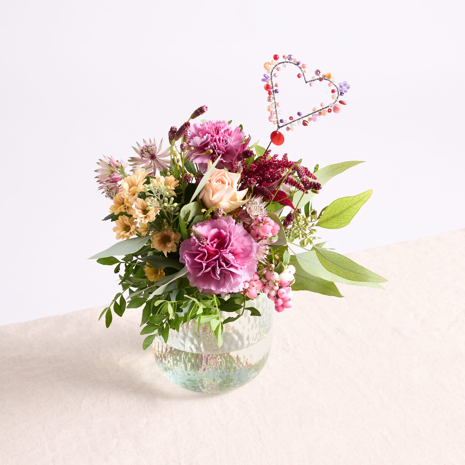 Ein farbenfroher Strauß rosa, weißer und violetter Blumen in einer Glasvase steht auf einer hellen Fläche vor einem schlichten Hintergrund.