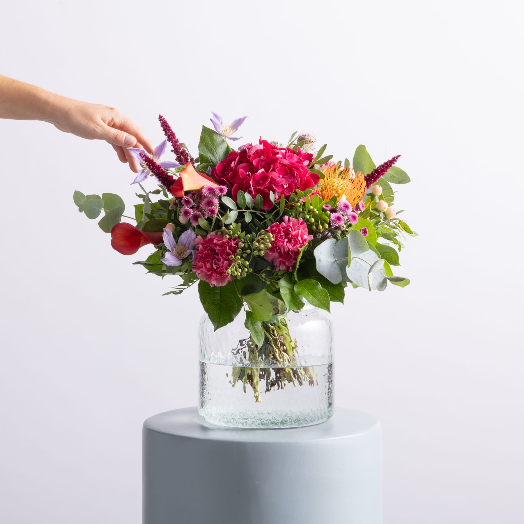 Ein leuchtender Strauß bunter Blumen, darunter rote Rosen, rosa Nelken und grünes Laub, steht in einer Glasvase vor einem schlichten weißen Hintergrund.