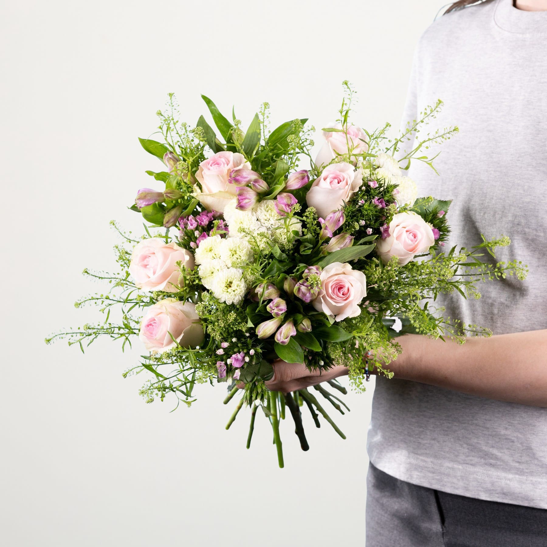 Ein leuchtender Strauß aus rosa Rosen, weißen Chrysanthemen und üppigem Grün wird vor einem schlichten weißen Hintergrund in den Händen einer Person gehalten.