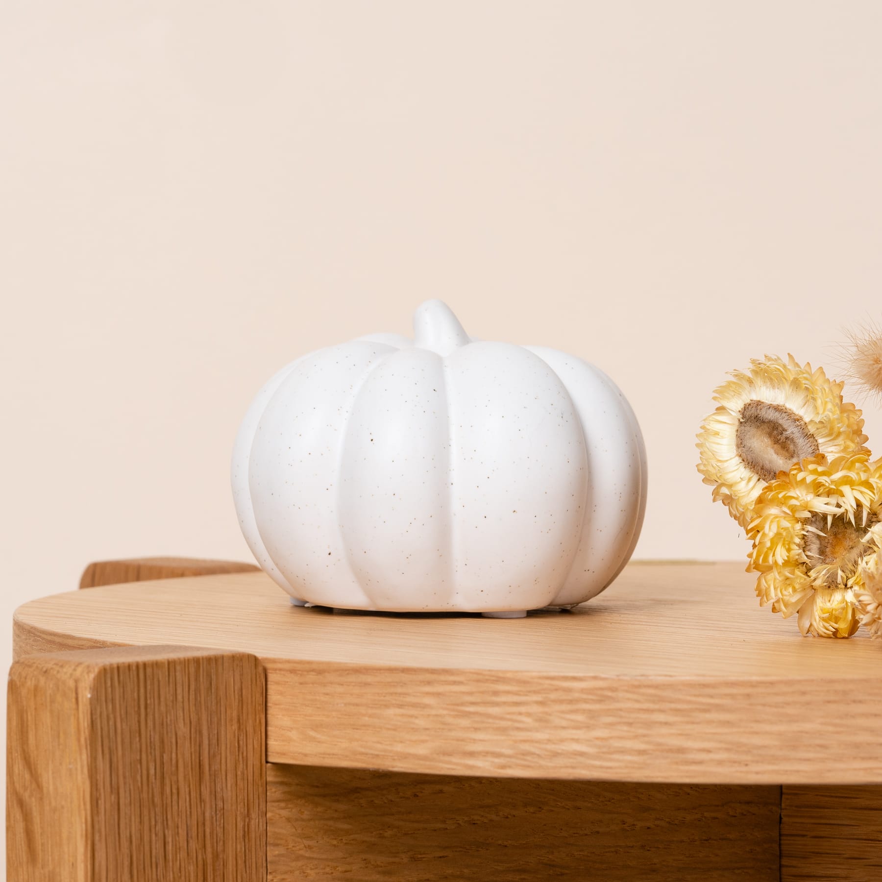 Ein weißer Keramikkürbis steht auf einer Holzoberfläche, im Hintergrund ist eine Sonnenblume zu sehen.