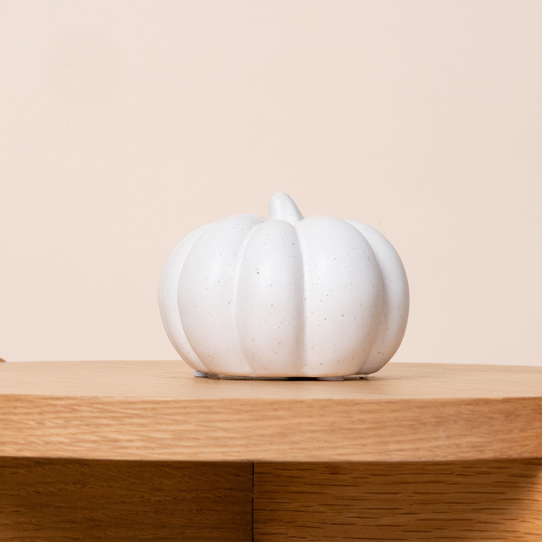 Ein weißes, kürbisförmiges Keramikobjekt liegt auf einer Holzoberfläche vor einem hellen Hintergrund.