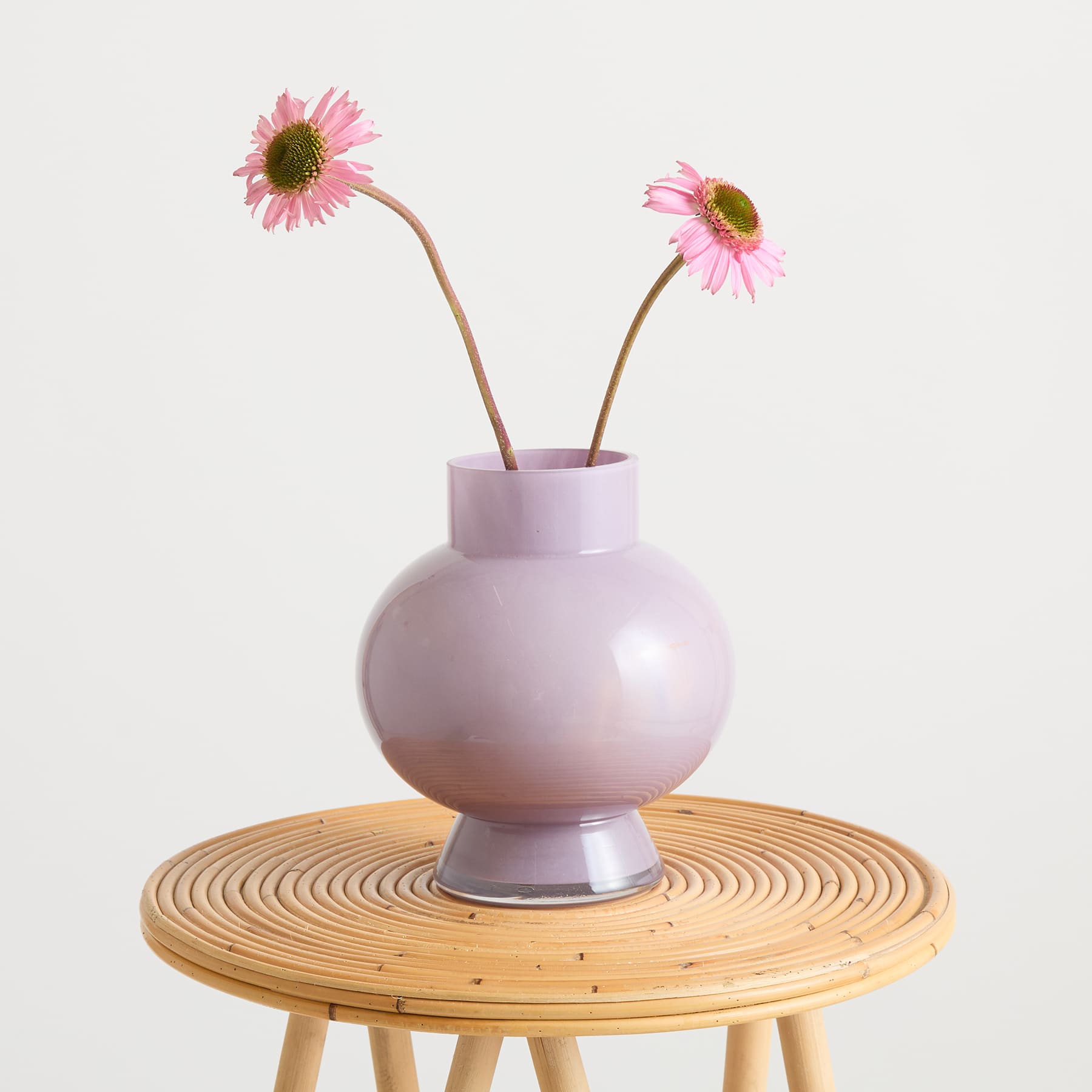 Eine pastellfarbene Vase mit zwei leuchtend rosa Gerbera-Blumen steht auf einem runden Holztisch vor einem schlichten weißen Hintergrund.