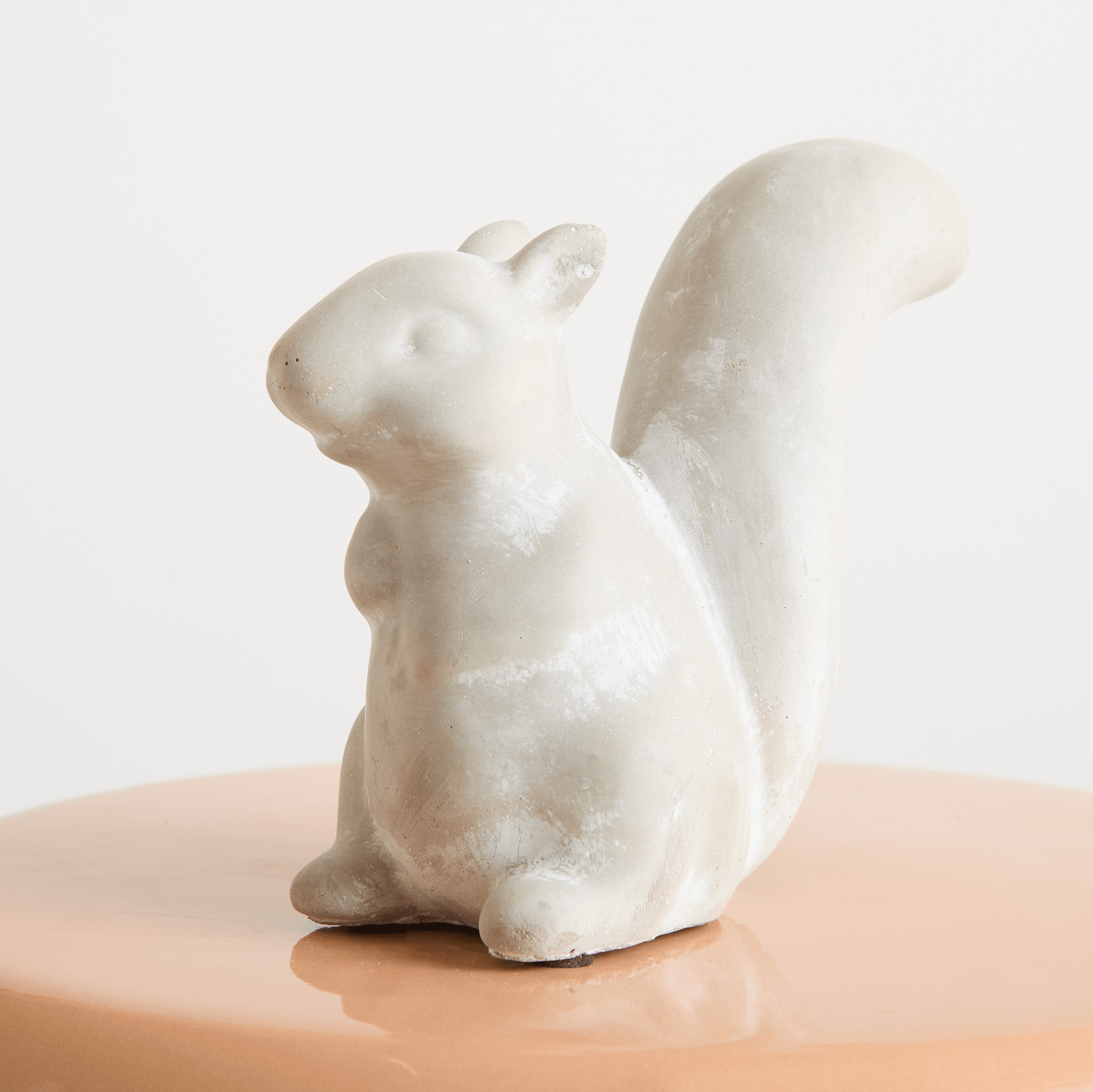 Eine kleine weiße Keramikfigur eines Eichhörnchens sitzt auf einer Holzoberfläche vor einem schlichten, hellen Hintergrund.