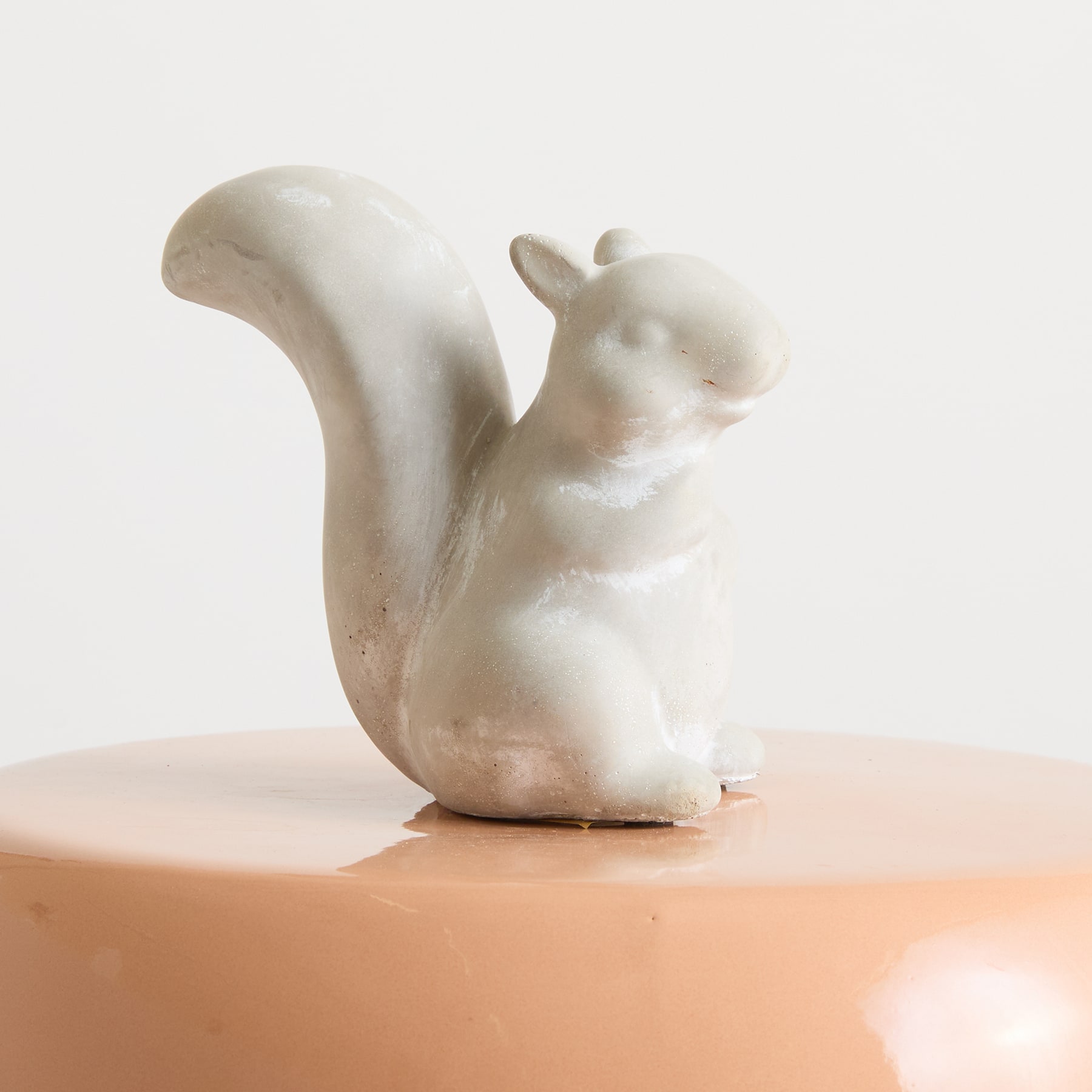 Eine weiße Keramikfigur eines Eichhörnchens sitzt auf einer orangefarbenen Oberfläche vor einem schlichten weißen Hintergrund.