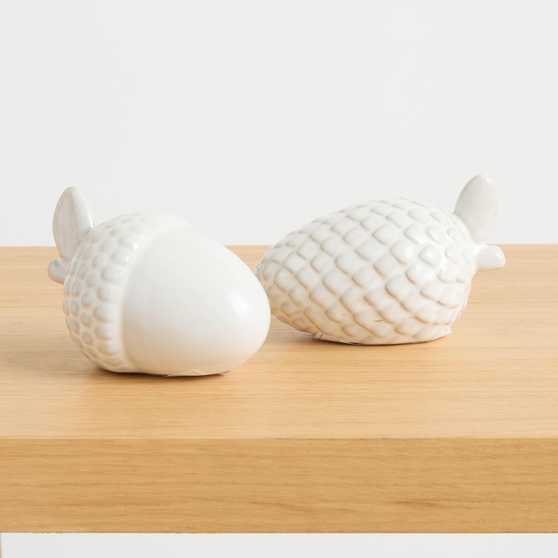 Zwei weiße Keramikobjekte, die Fischen oder Meerestieren ähneln, sind auf einer Holzoberfläche vor einem schlichten weißen Hintergrund platziert.