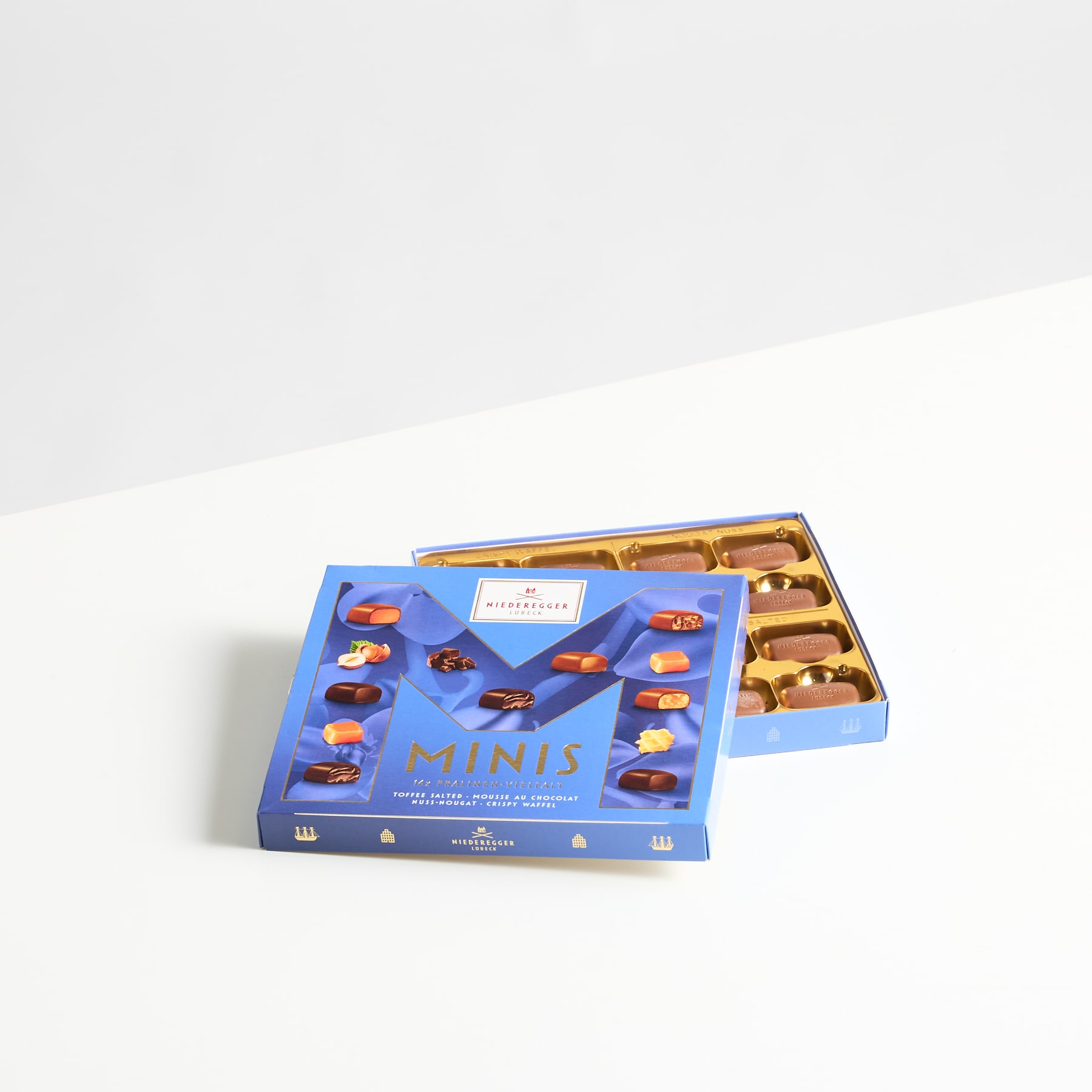 Auf einer weißen Fläche steht eine blaue Schachtel mit verschiedenen Miniatur-Schokoladenbonbons, daneben liegt ein goldfarbenes Utensil.