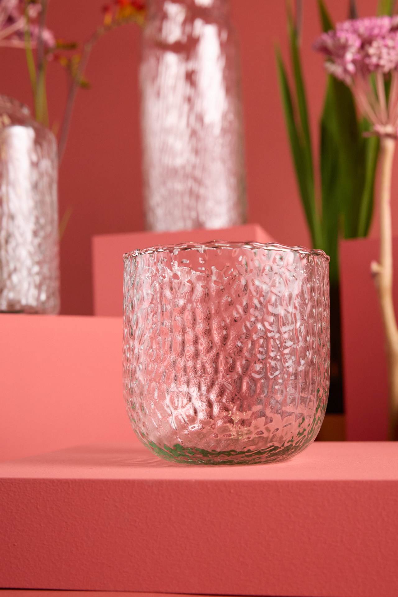 Eine rosa Glasschale mit strukturierter, gehämmerter Oberfläche steht auf einem rosa Hintergrund, umgeben von rosa Blumen.
