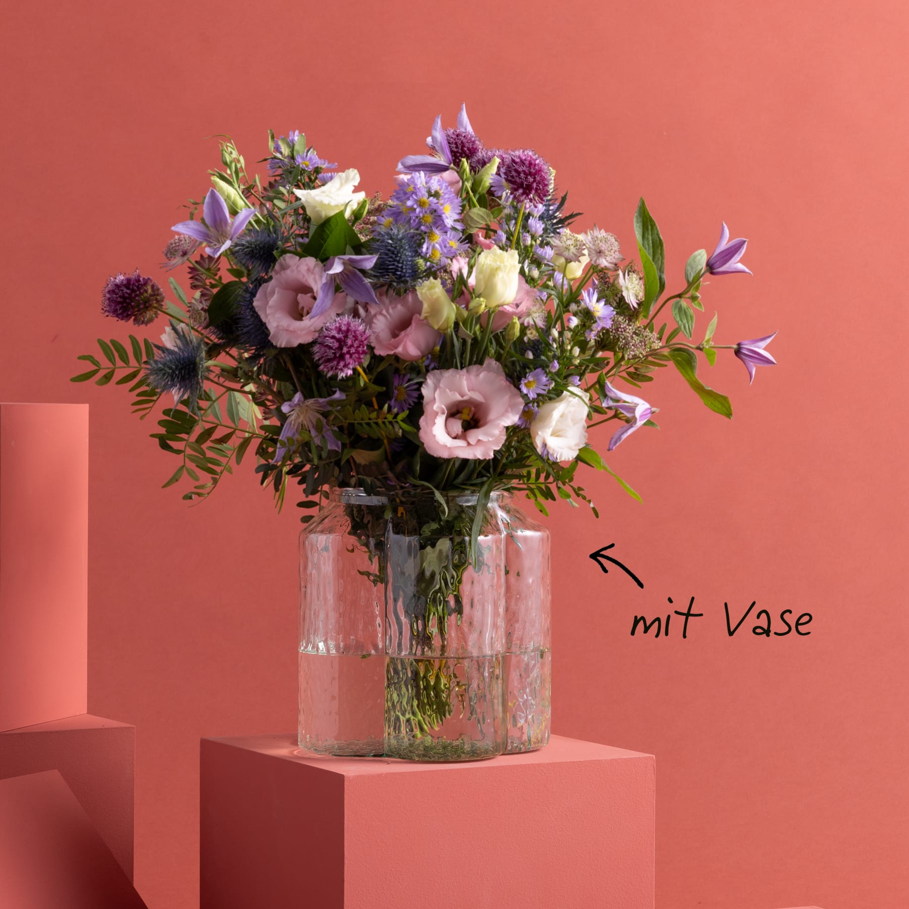 Ein farbenfroher Strauß verschiedener Blumen, darunter Rosen, Nelken und andere Blüten, wird in einer Glasvase vor einem warmen, korallenfarbenen Hintergrund präsentiert.