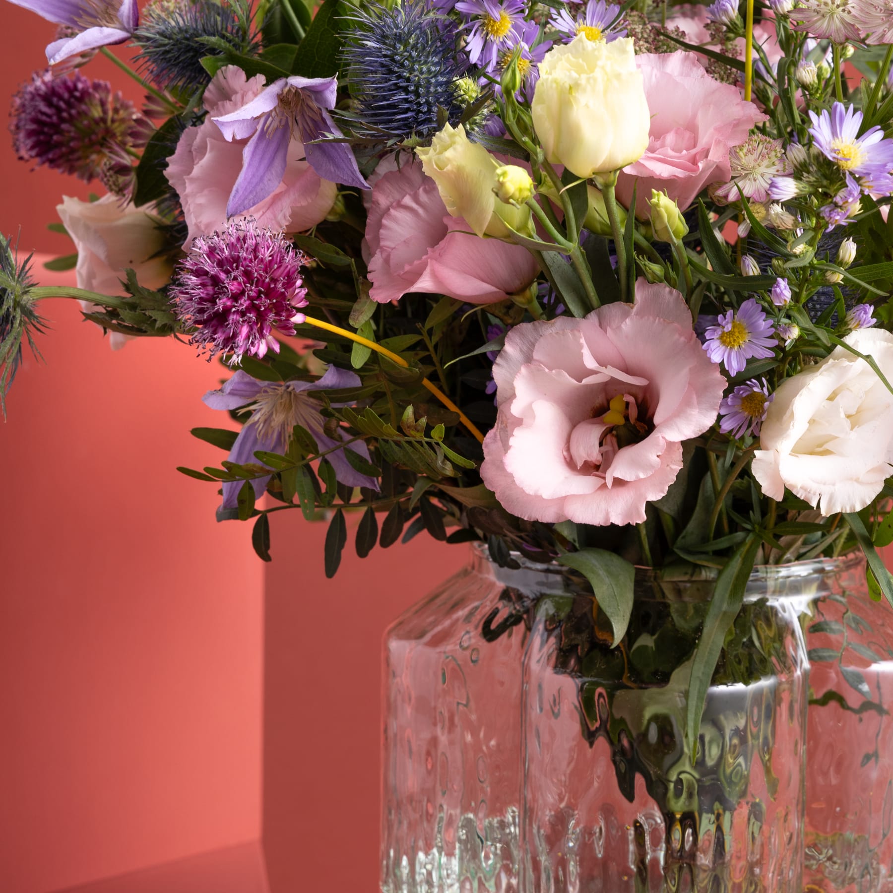 Ein farbenfroher Strauß verschiedener Blumen, darunter rosa Pfingstrosen, violette Disteln und gelbe Gänseblümchen, arrangiert in einer Glasvase vor einem warmen, orangeroten Hintergrund.