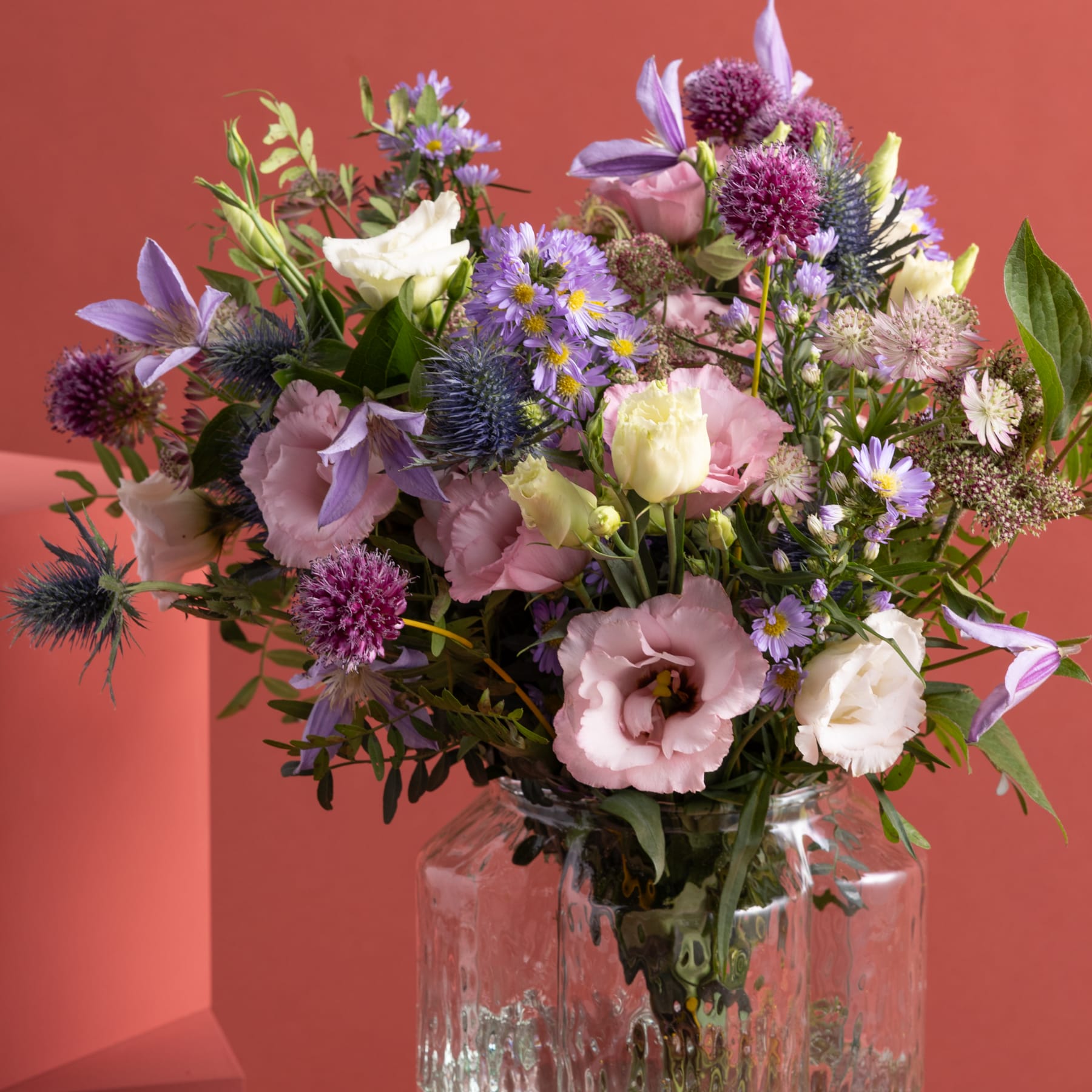 Ein farbenfroher Strauß verschiedener Blumen, darunter Rosen, Gänseblümchen und Disteln, wird in einer Glasvase vor einem warmen, rot-orangen Hintergrund präsentiert.