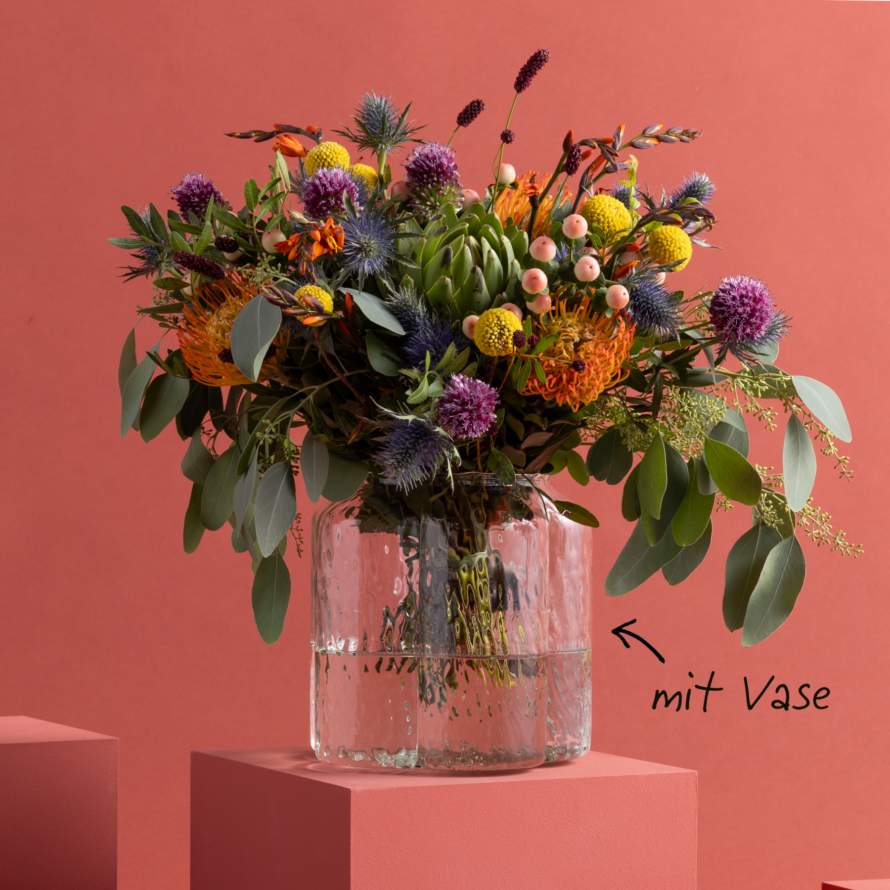 Ein farbenfroher Strauß herbstlich gefärbter Blumen, darunter Artischocken, Dahlien und andere üppige Blüten, wird in einer Glasvase vor einem kräftigen korallenfarbenen Hintergrund präsentiert.