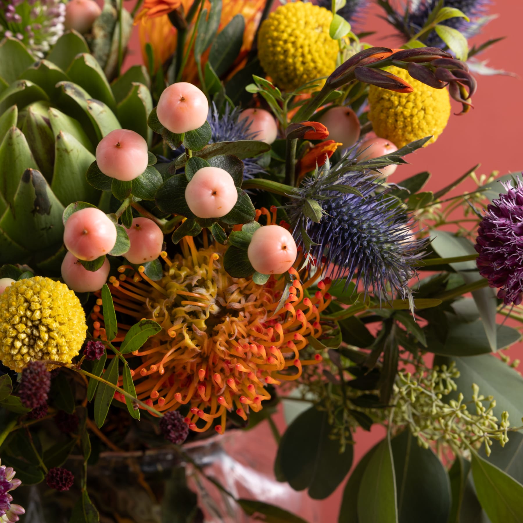 Ein lebendiger Strauß bunter Blumen, darunter gelbe Chrysanthemen, rosa Beeren und eine auffällige violette Distel, vor einem zartrosa Hintergrund.