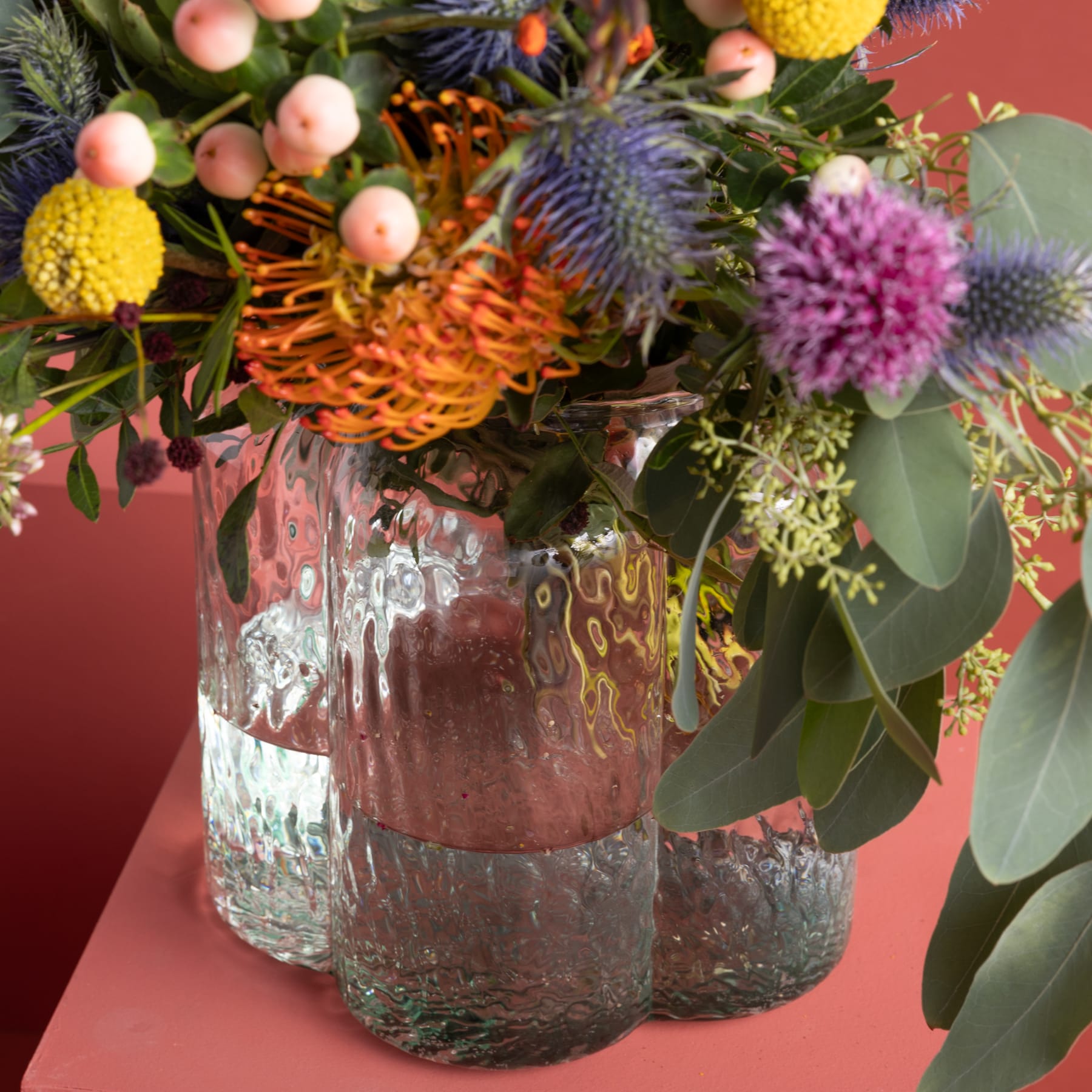 Ein lebendiger Strauß bunter Blumen, darunter orangefarbene Proteas, violette Disteln und gelbe, pomponartige Blüten, arrangiert in einer strukturierten Glasvase vor einem tiefroten Hintergrund.