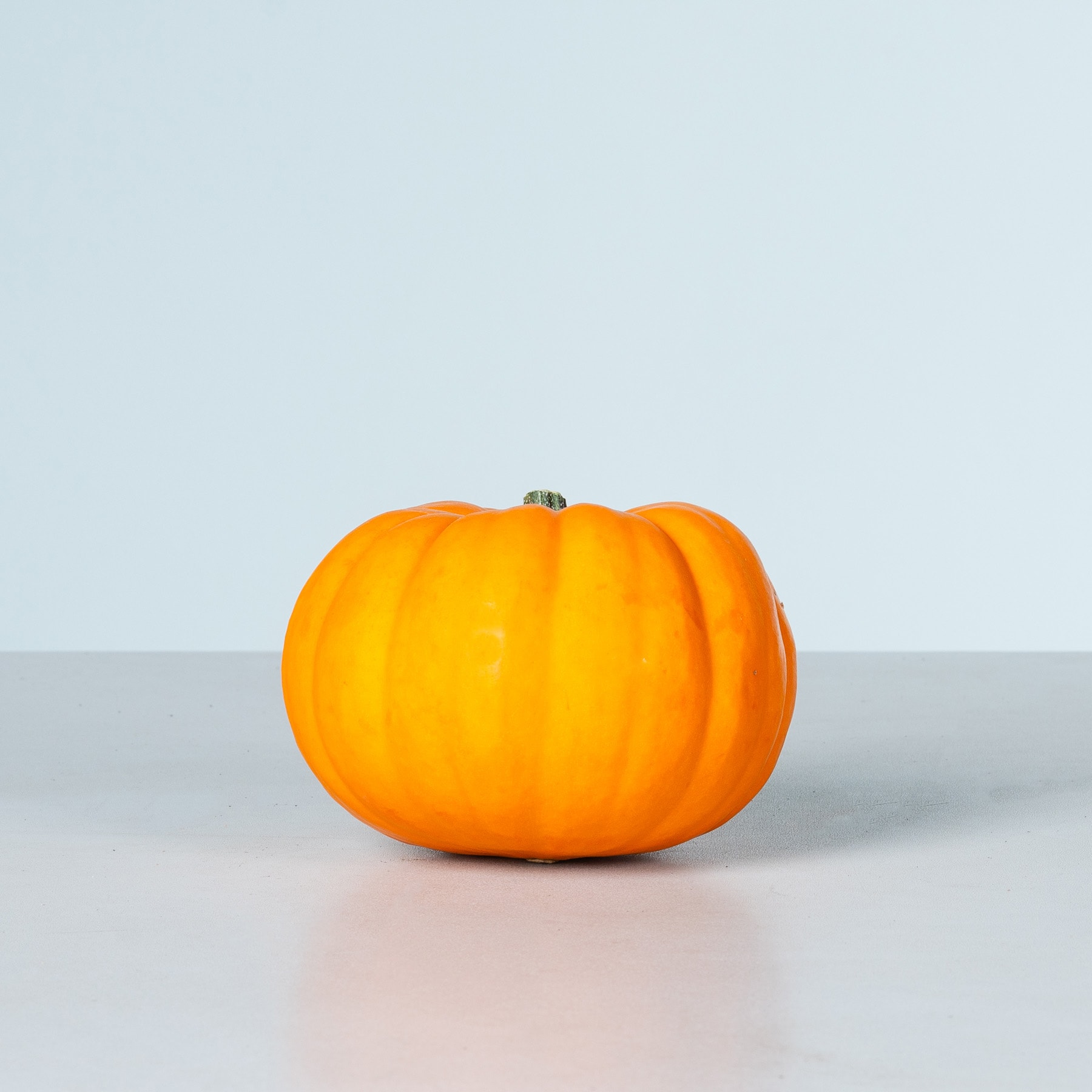 Ein leuchtend orangefarbener Kürbis liegt auf einer schlichten, hellen Oberfläche vor einem hellblauen Hintergrund.