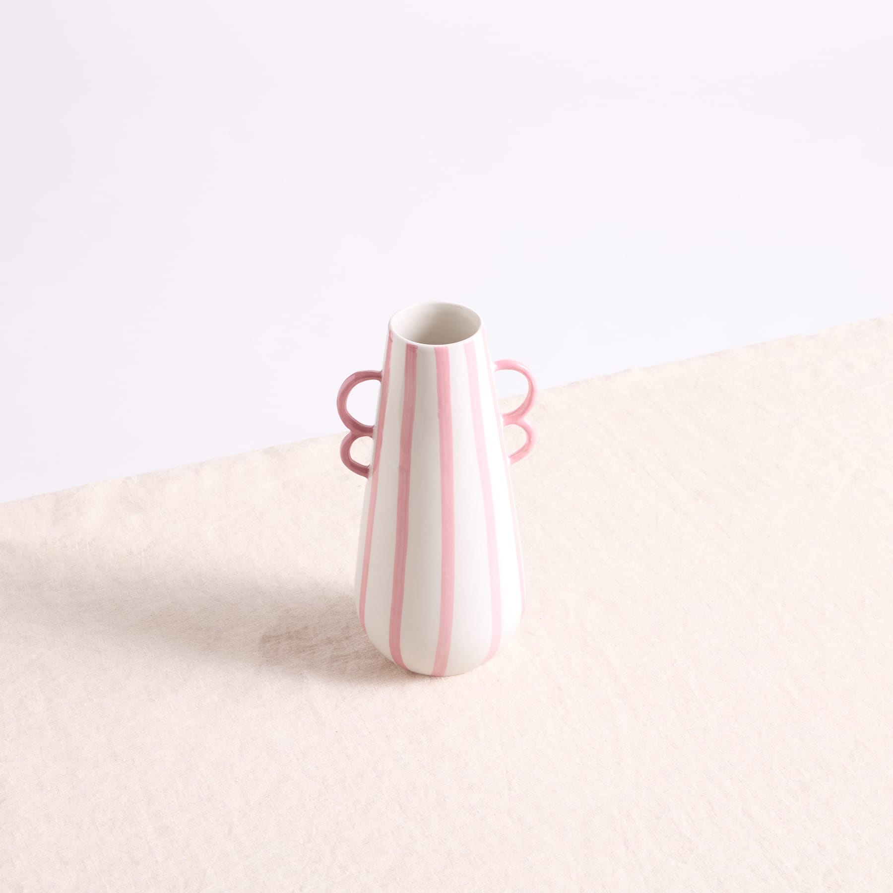 Eine weiße Keramikvase mit rosa Streifen steht auf einer schlichten, hellen Oberfläche vor einem schlichten weißen Hintergrund.