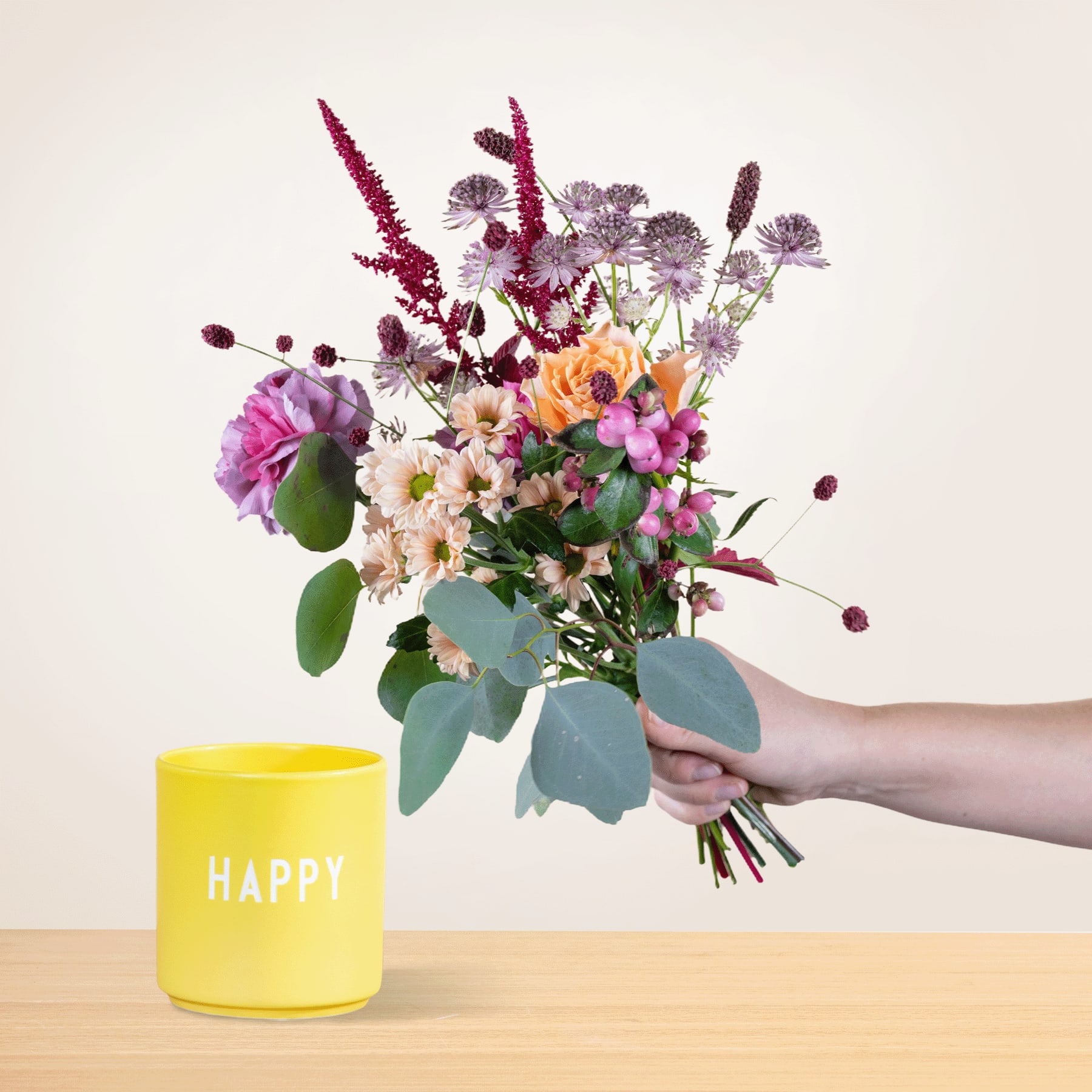 Ein leuchtender Strauß bunter Blumen, darunter rosa, violette und gelbe Blüten, wird in der Hand einer Person gehalten. Vor einem gelben Behälter steht auf einem schlichten Hintergrund das Wort „HAPPY“.