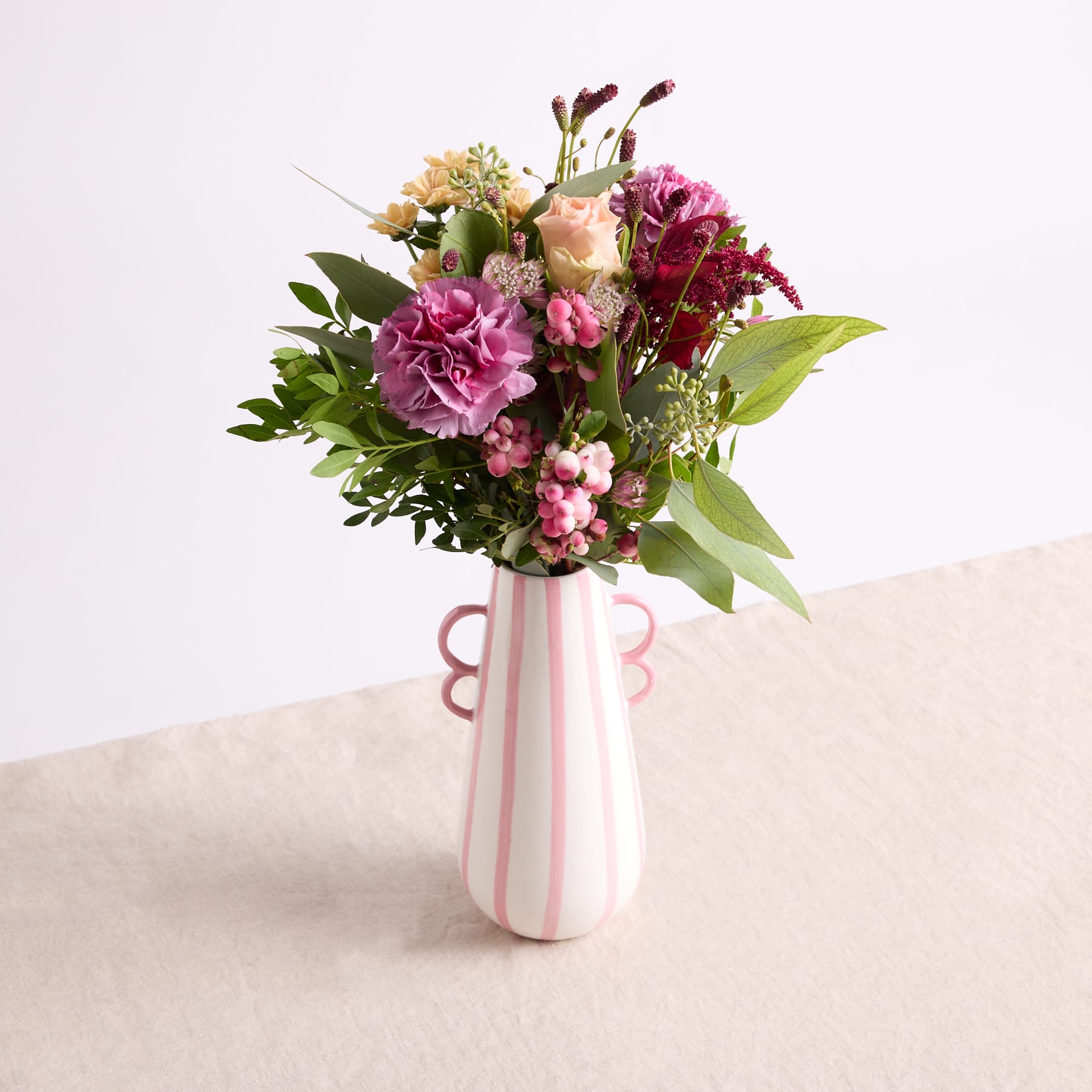 Ein farbenfroher Strauß rosa und weißer Blumen, darunter Pfingstrosen und Nelken, wird in einer rosa und weiß gestreiften Vase vor einem schlichten Hintergrund präsentiert.