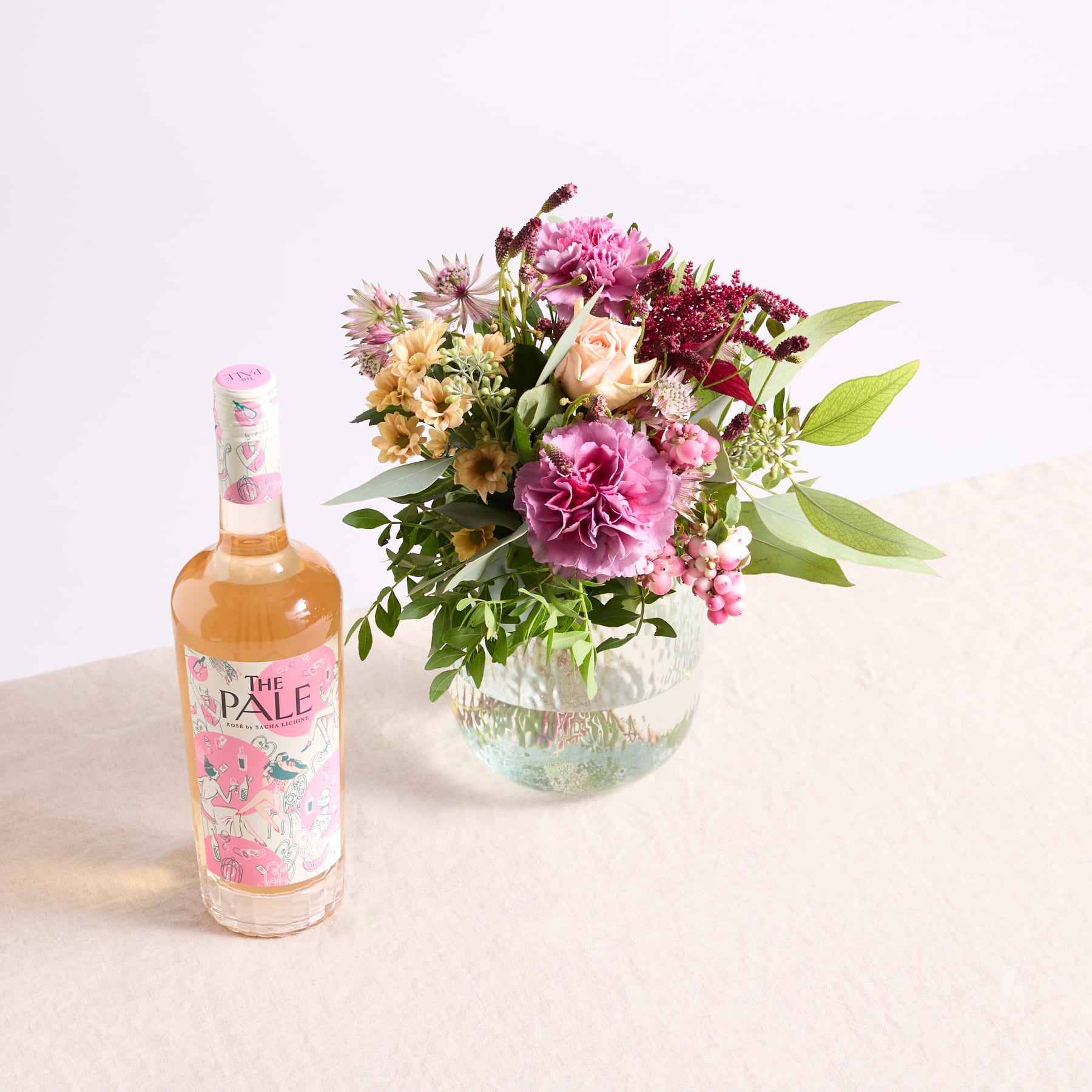 Ein leuchtender Strauß rosa, weißer und violetter Blumen in einer Glasvase steht neben einer Flasche hellem Roséwein auf einer hellen Oberfläche.