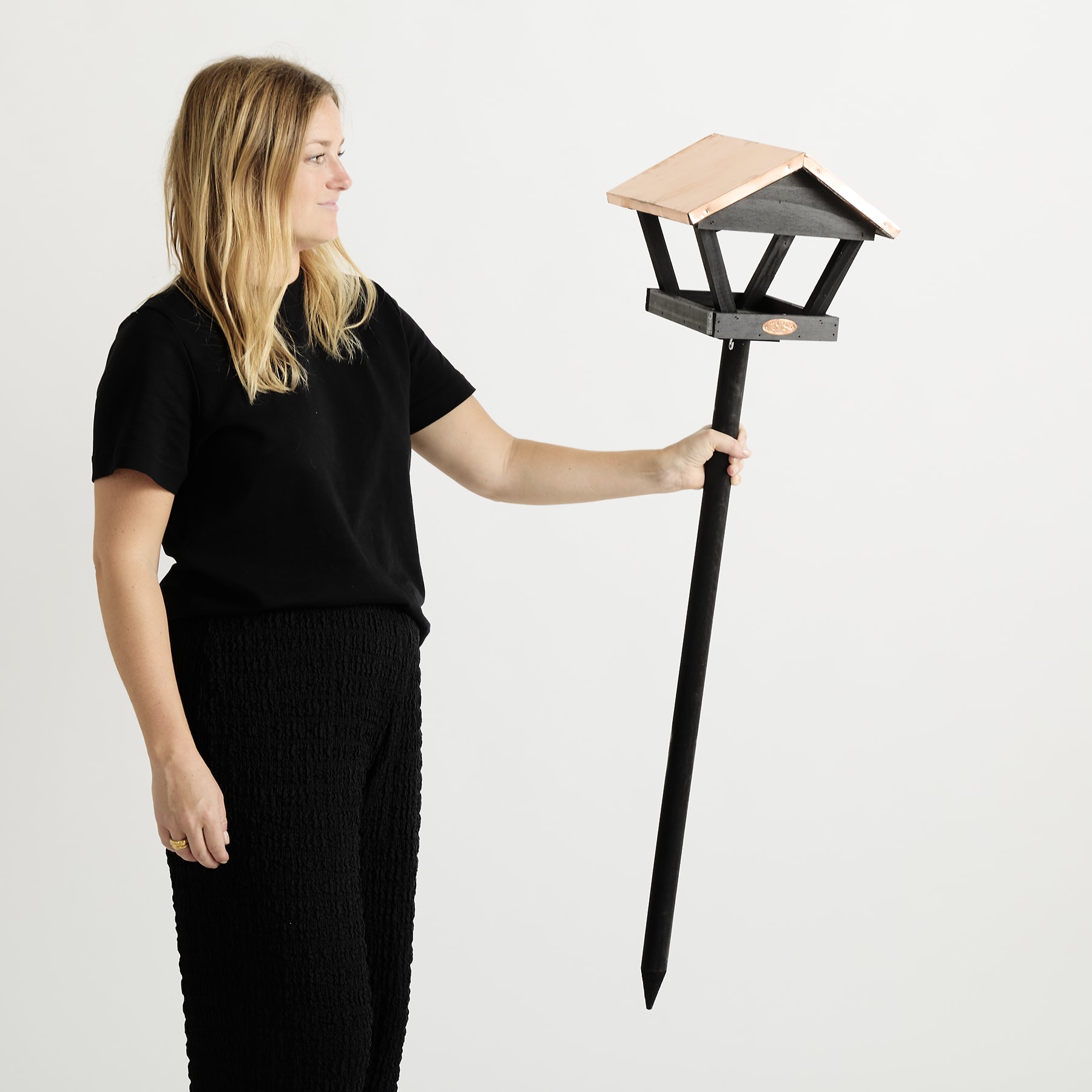 Eine junge Frau in einem schwarzen Outfit steht vor einem schlichten weißen Hintergrund und hält eine dekorative Außenleuchte in der Hand.