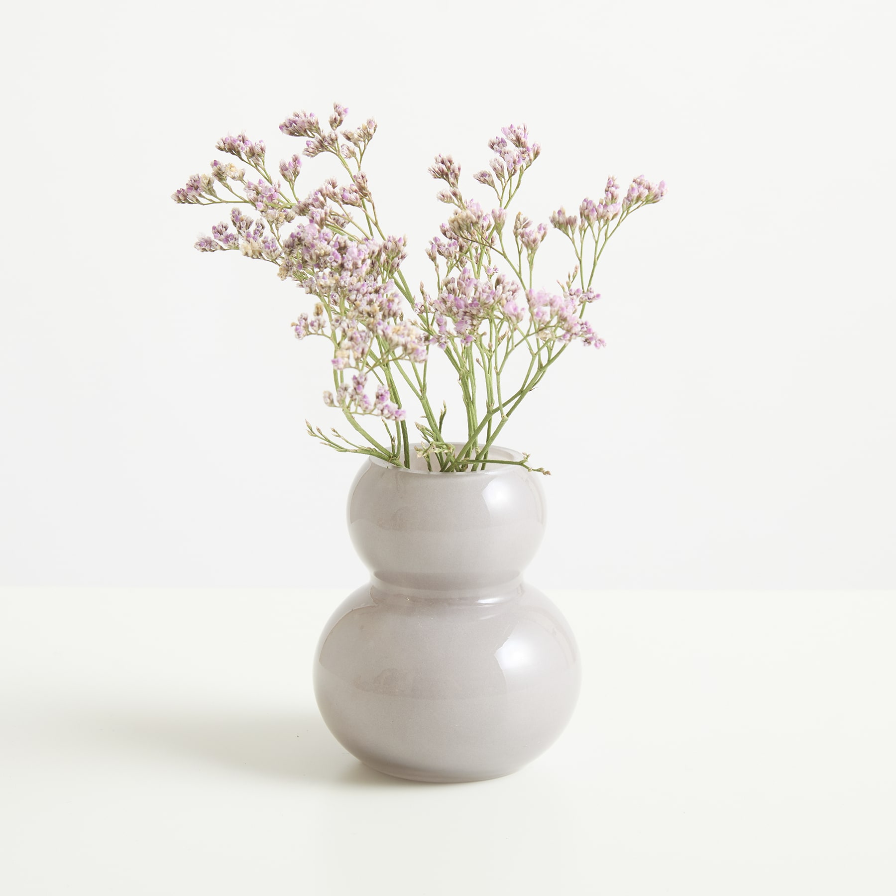 Eine weiße Keramikvase mit einem Strauß zarter, blassrosa Blumen steht vor einem schlichten, weißen Hintergrund.
