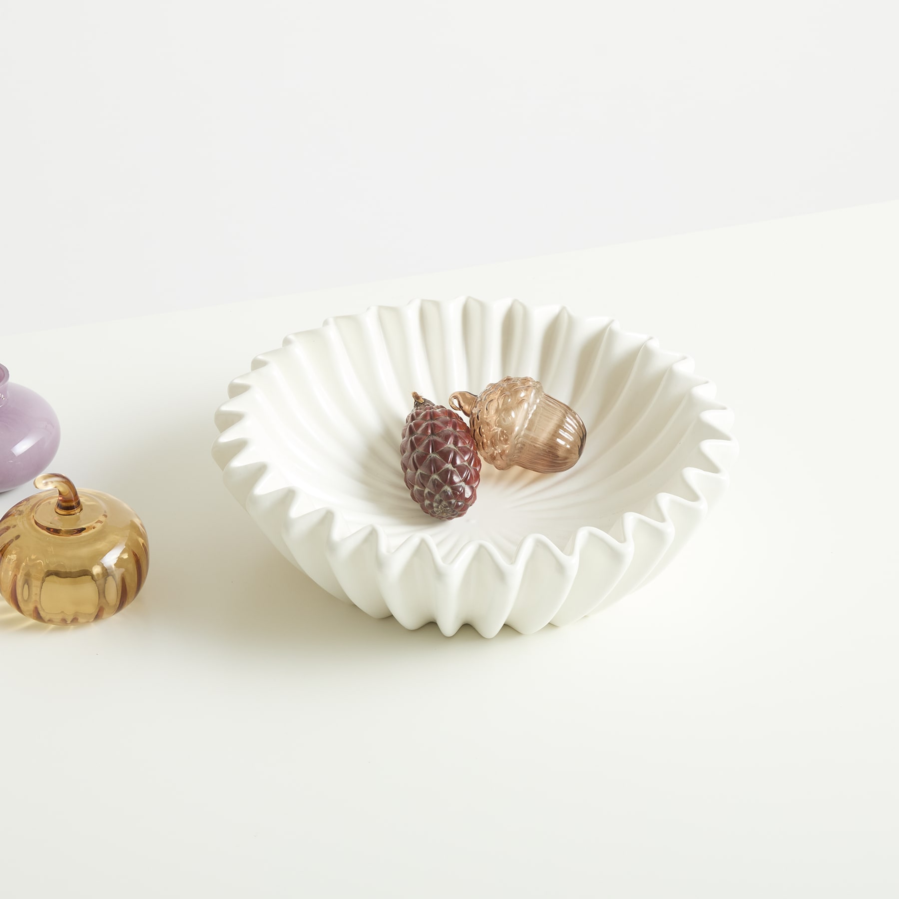 Eine weiße Keramikschale mit gewelltem Rand enthält einen Tannenzapfen und ein kleines Glasornament auf einem schlichten weißen Hintergrund.