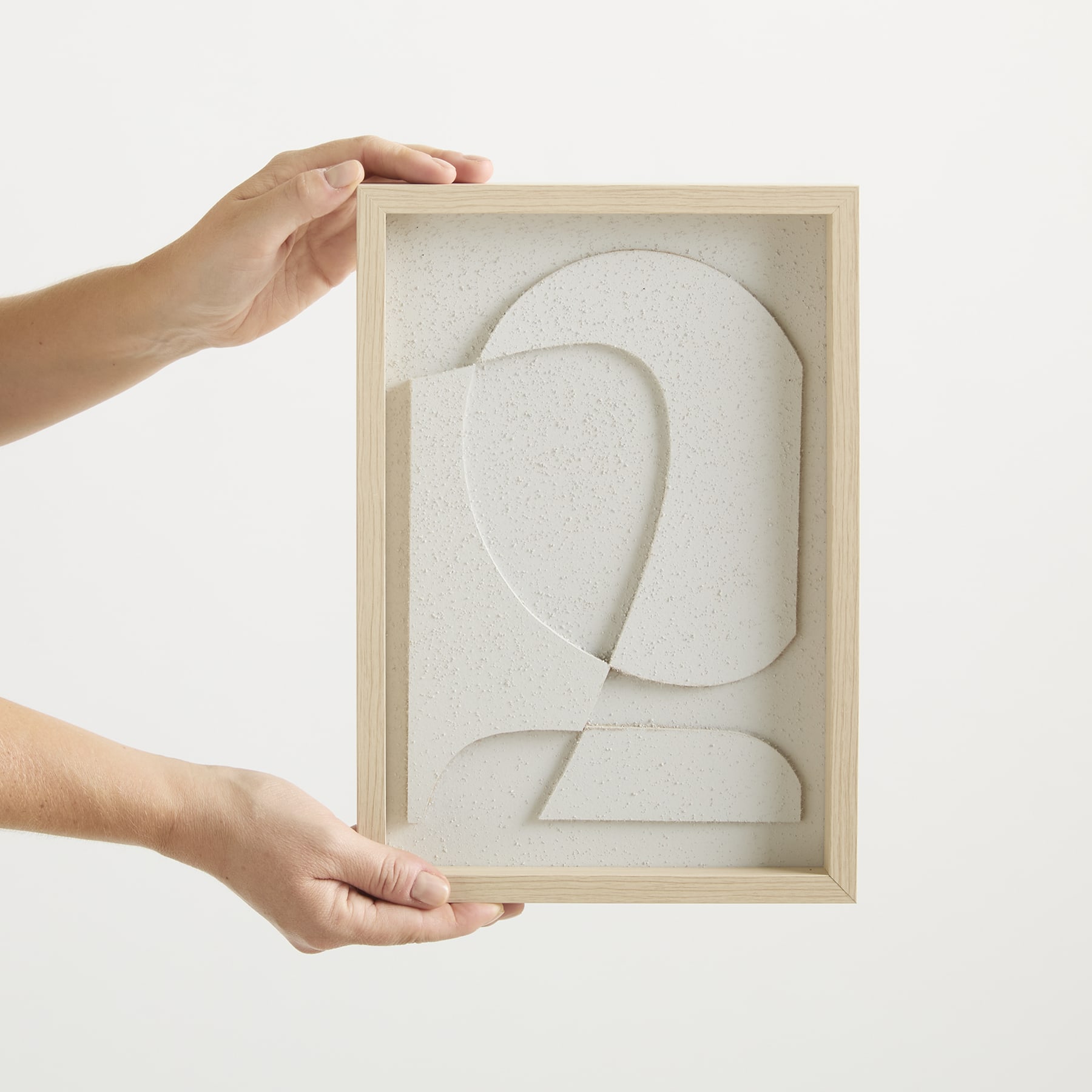 Ein Holzrahmen, der eine weiße, abstrakte Skulptur mit geschwungenen und überlappenden Formen enthält, wird von den Händen einer Person gehalten.