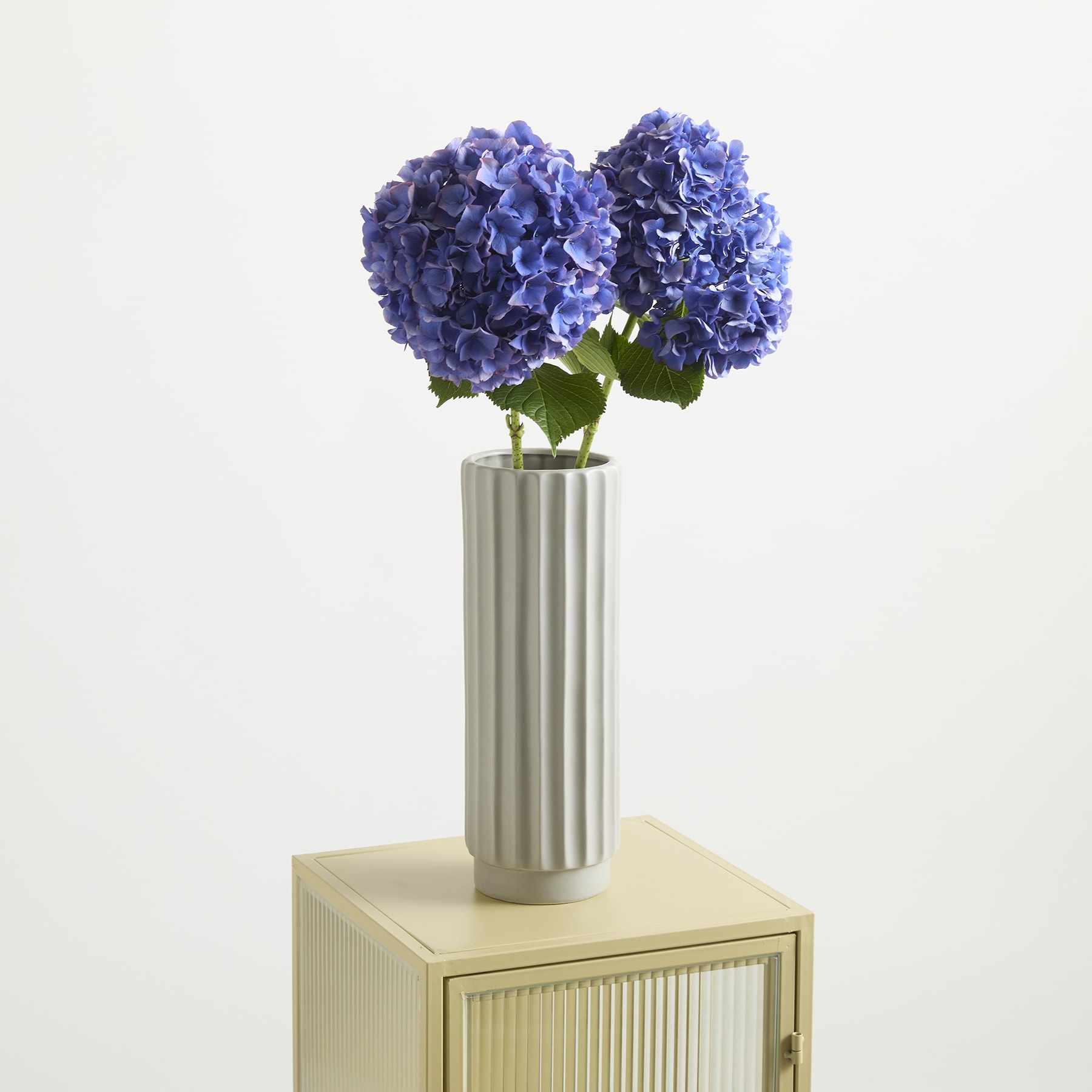 Eine Vase mit leuchtend violetten Hortensienblüten steht auf einer Holzoberfläche vor einem schlichten weißen Hintergrund.