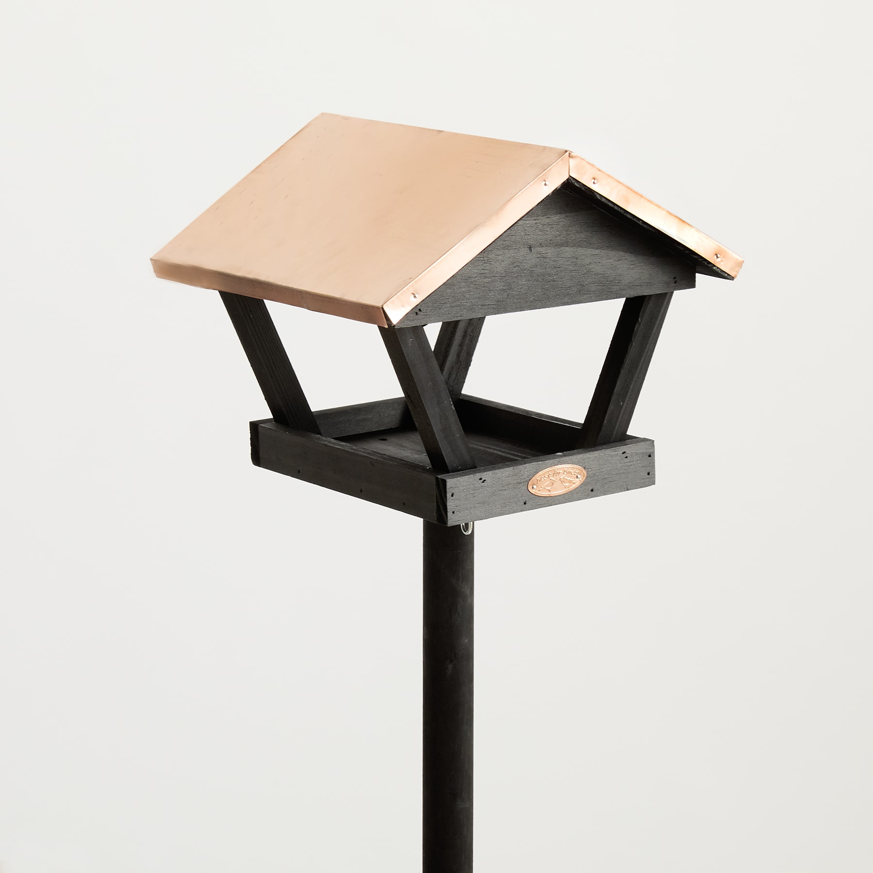 Ein hölzernes Vogelhäuschen mit Spitzdach steht auf einer schwarzen Metallstange vor einem schlichten weißen Hintergrund.