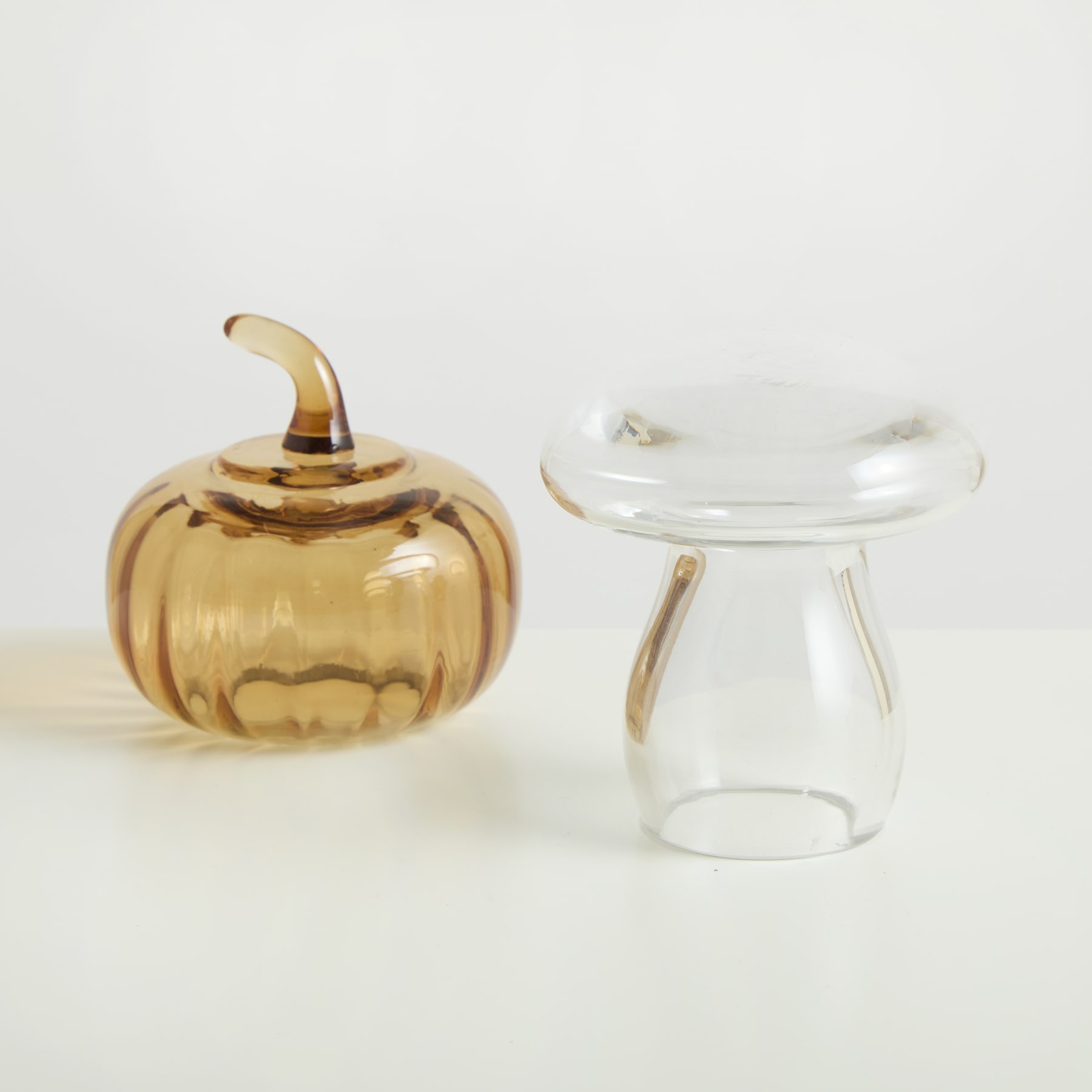 Zwei Glasbehälter, einer davon ein goldfarbenes, kürbisförmiges Gefäß mit Deckel und der andere ein klares, abgerundetes Glasgefäß mit Deckel, stehen auf einem schlichten weißen Hintergrund.