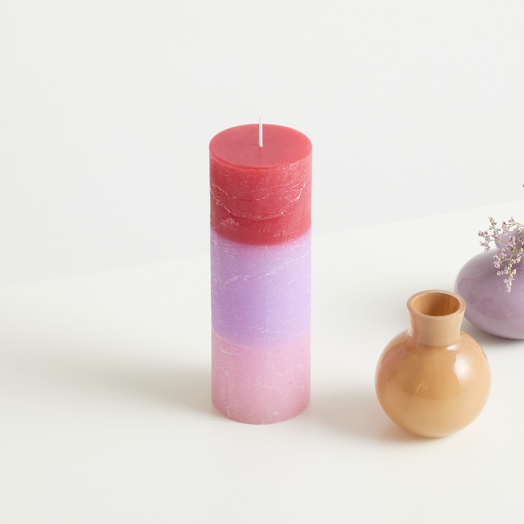 Eine rot-rosa Kerze steht neben einer kleinen gelben Vase auf einem schlichten weißen Hintergrund.