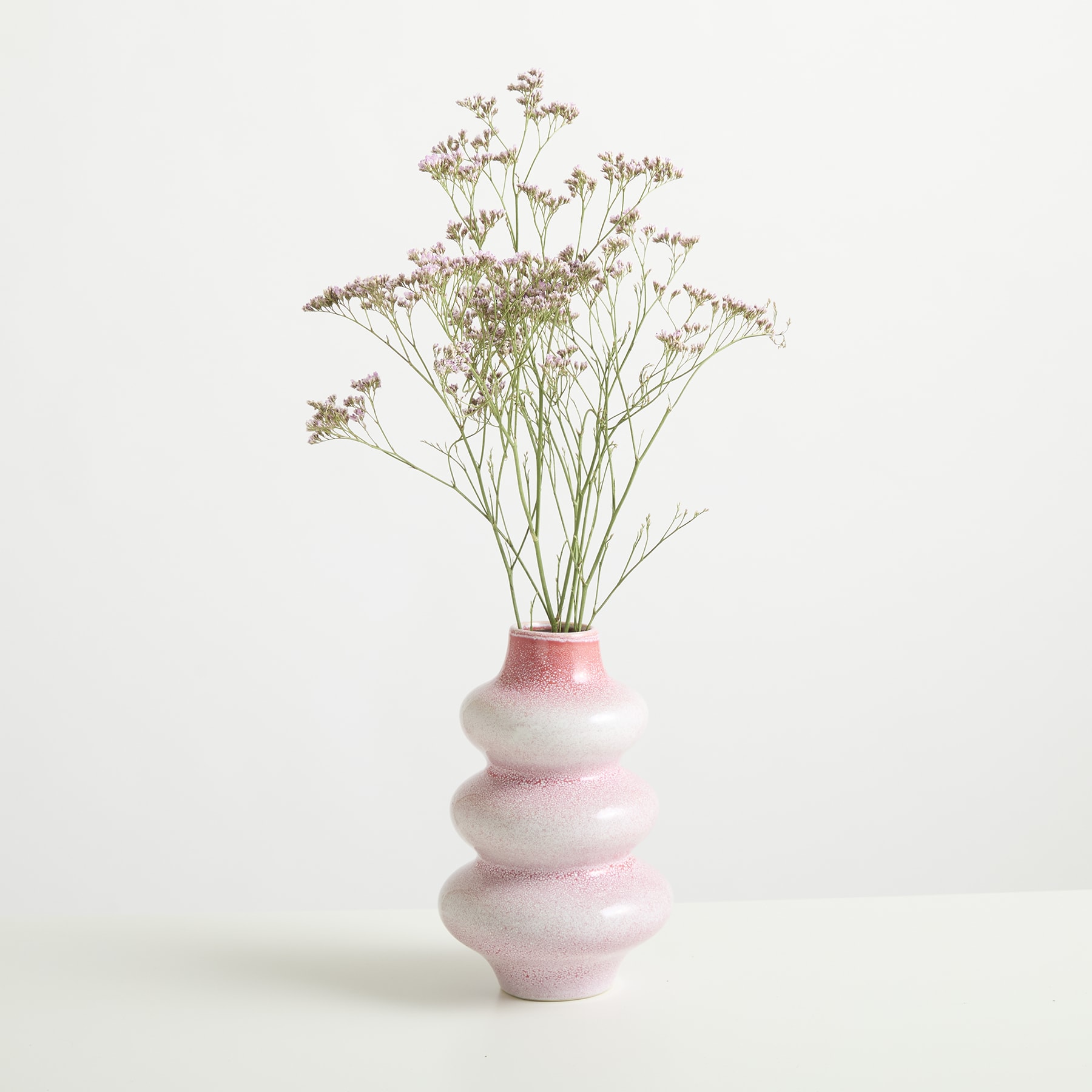 Ein zarter Strauß kleiner weißer Blumen wird in einer minimalistischen rosa-weißen Keramikvase vor einem schlichten weißen Hintergrund präsentiert.