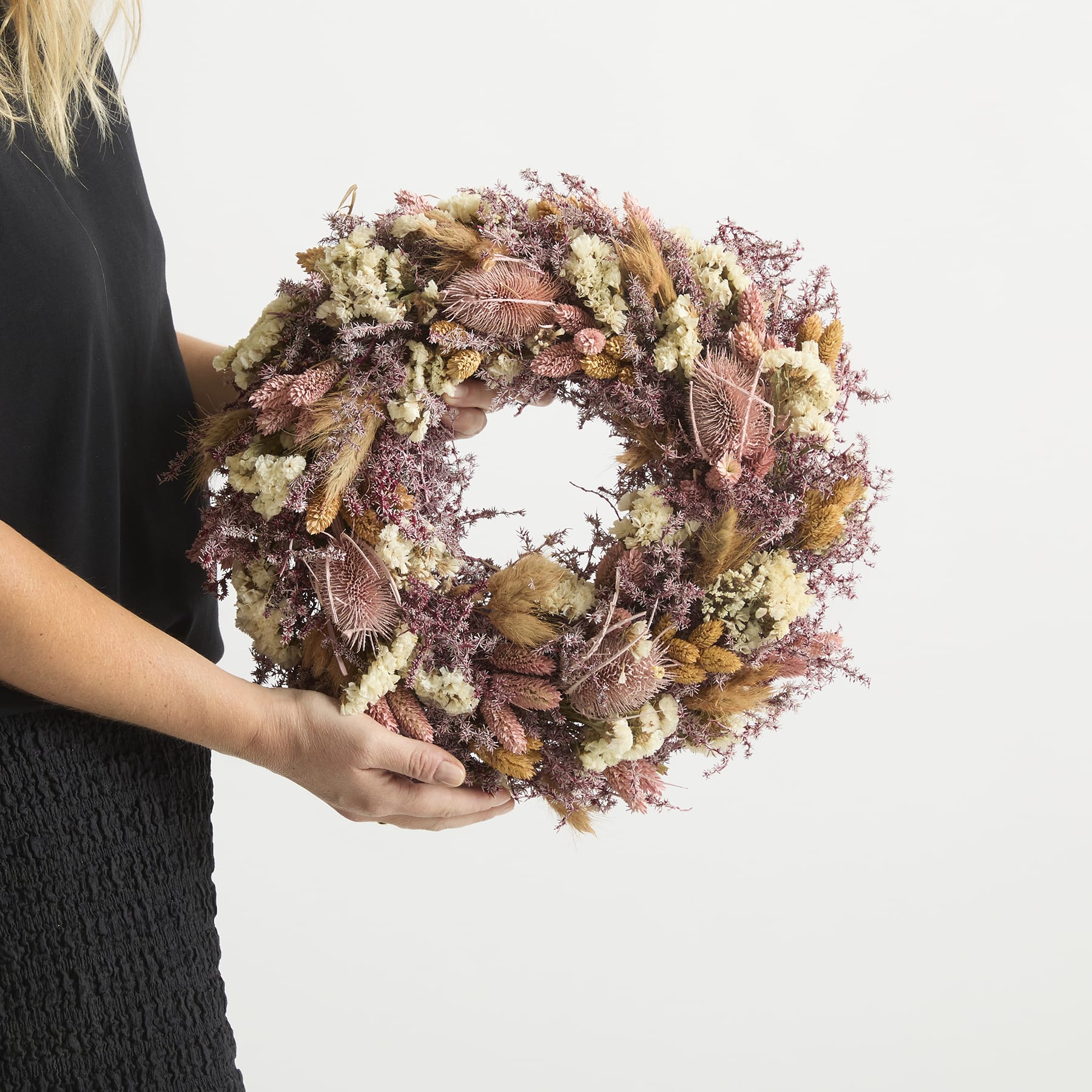 Ein farbenfroher Herbstkranz aus getrockneten Blumen, Blättern und anderen Naturelementen wird von einer Person vor einem schlichten weißen Hintergrund in den Händen gehalten.