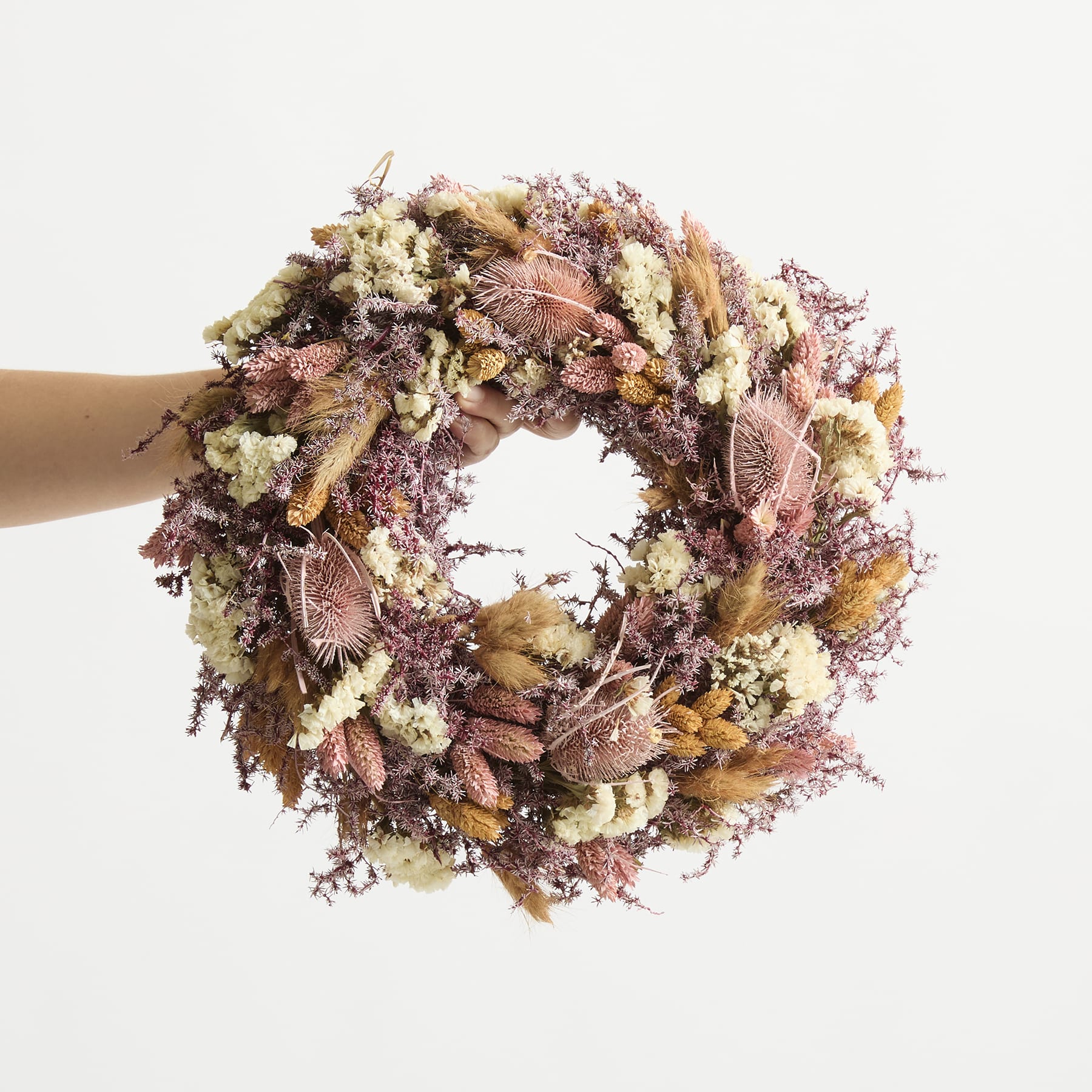 Ein farbenfroher, getrockneter Blumenkranz aus verschiedenen Herbstblättern, Blumen und anderen natürlichen Elementen wird vor einem schlichten weißen Hintergrund in der Hand einer Person gehalten.